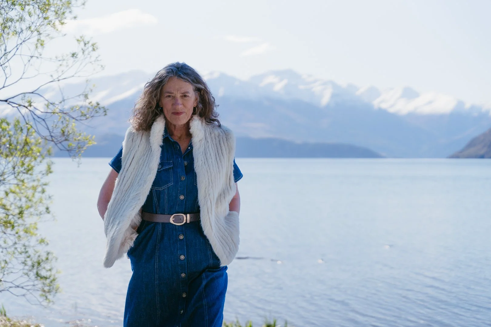 An older woman with curly gray hair outdoors by a lake, wearing a denim dress and a white fur vest, with snow-capped mountains in the background.