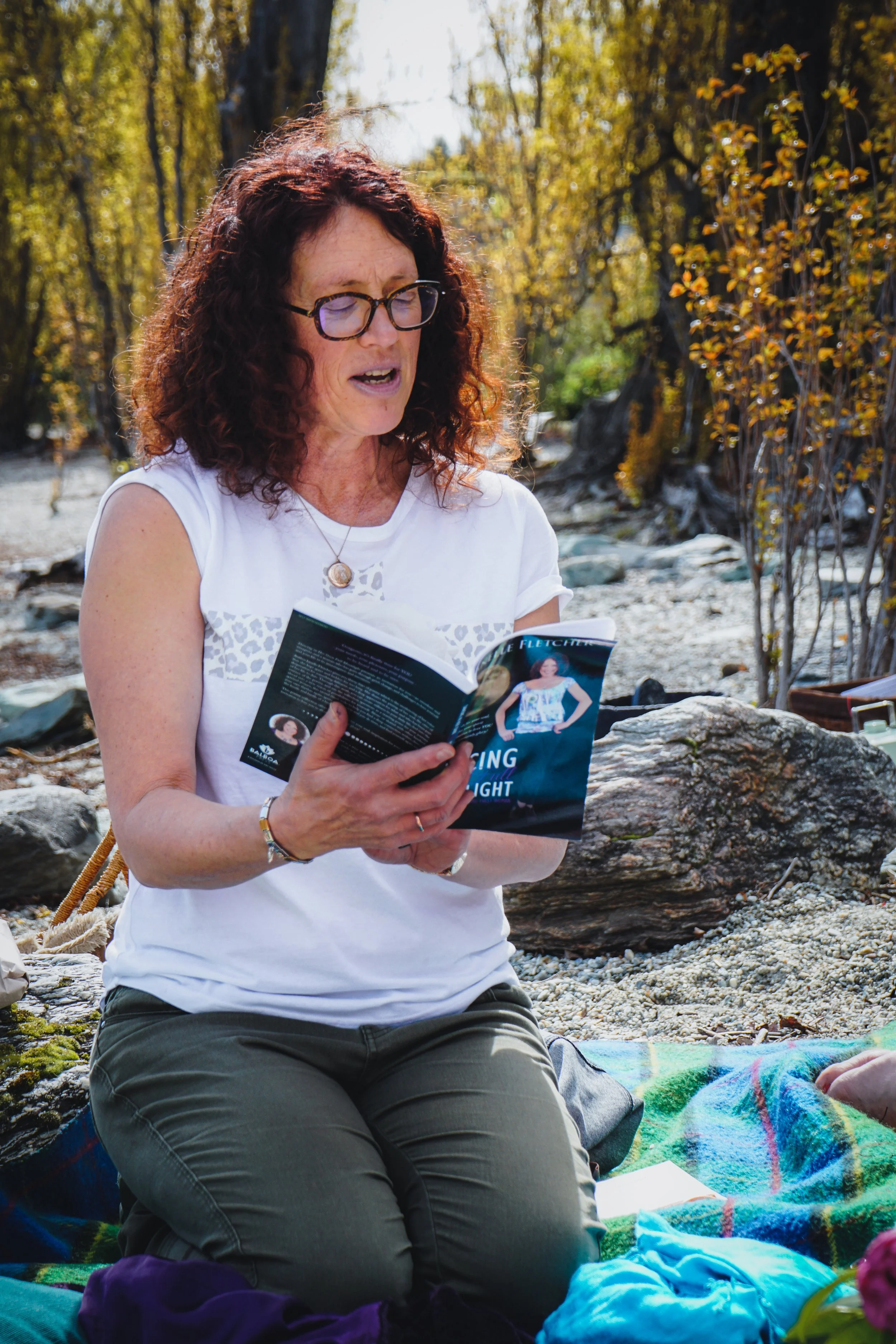 Woman with glasses reading a book outdoors on a rocky beach with trees and fallen leaves in the background.