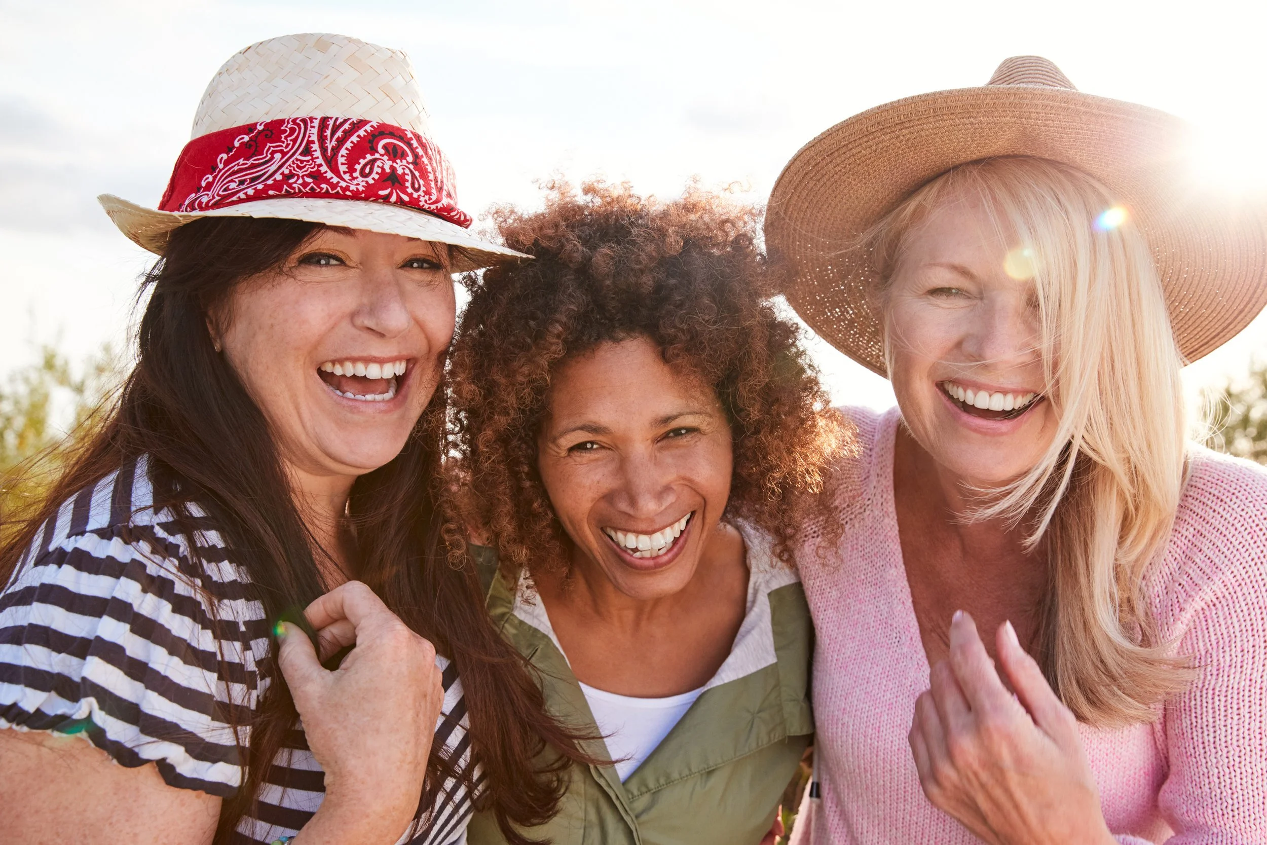 Three women outdoors smiling and enjoying time together, wearing hats and casual clothing, with sunlight in the background.