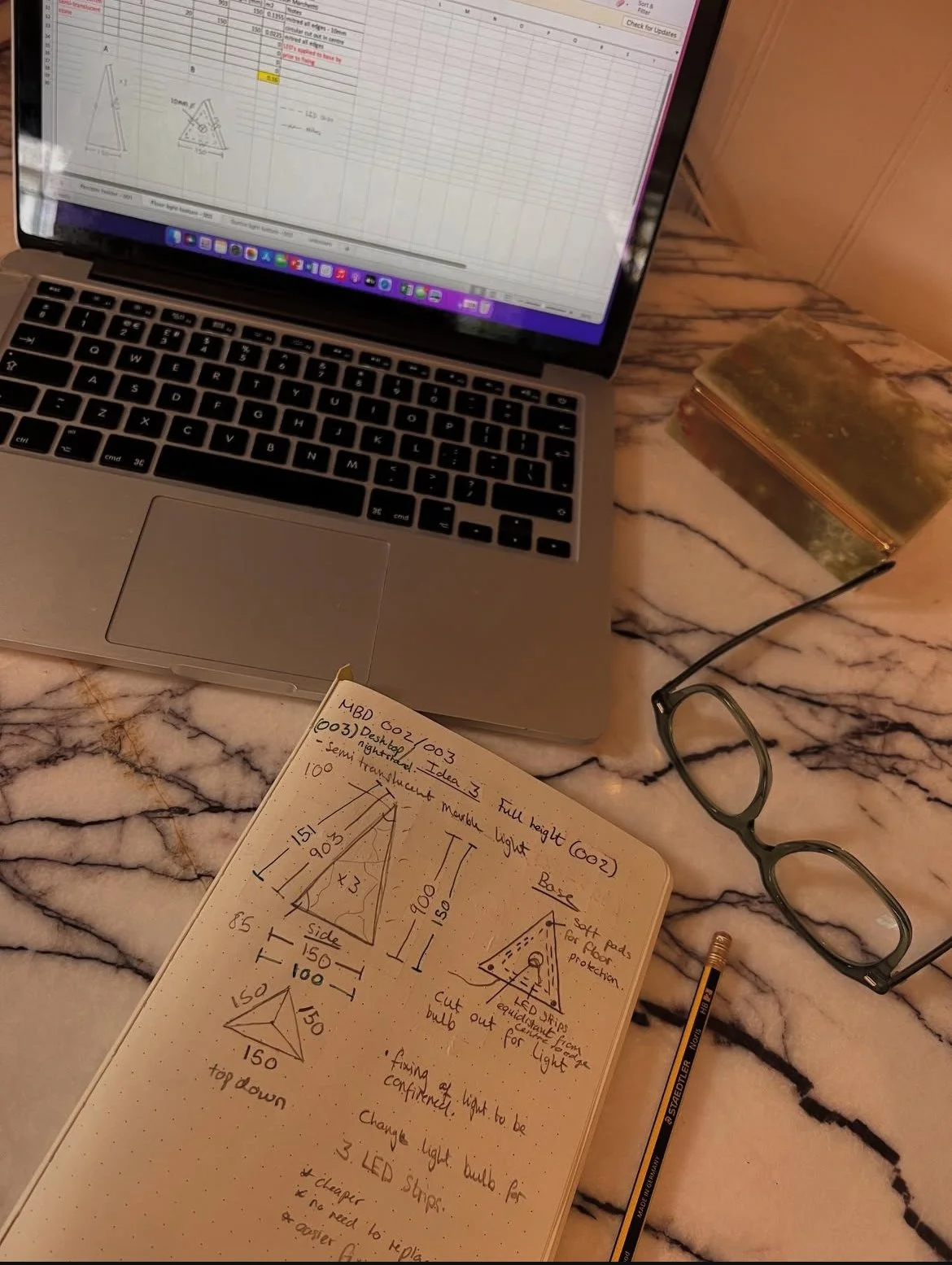 A workspace with a MacBook laptop displaying a spreadsheet, a pair of glasses, a notebook with sketches and notes on LED light design, a pencil, and a stone box on a marble or granite surface.