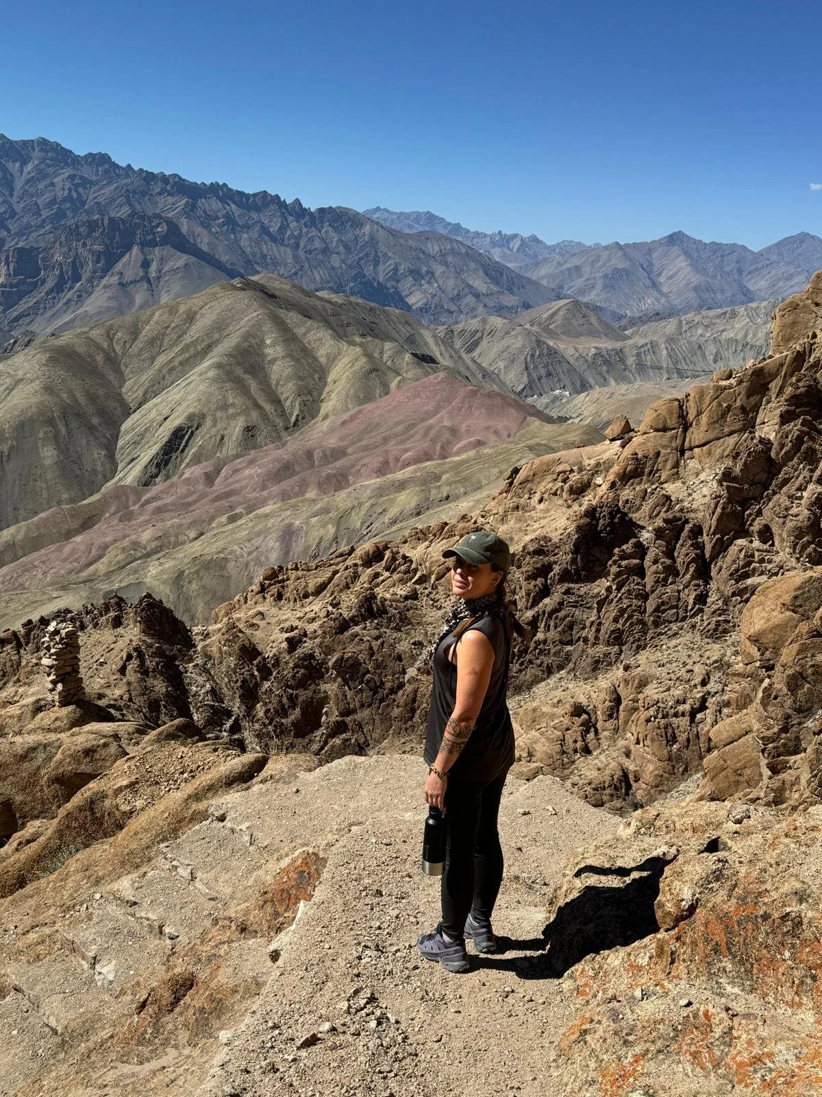Une femme en tenue de randonnée avec un chapeau et une gourde, se tenant sur un sentier rocheux dans un paysage de montagnes colorées et arides.