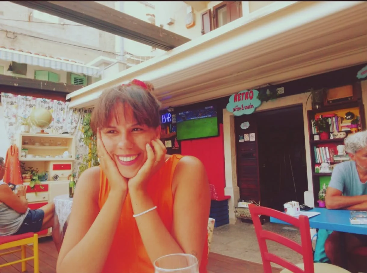 A woman smiling and resting her face in her hands at an outdoor café, with colorful decorations, a TV screen, books, and other people in the background.
