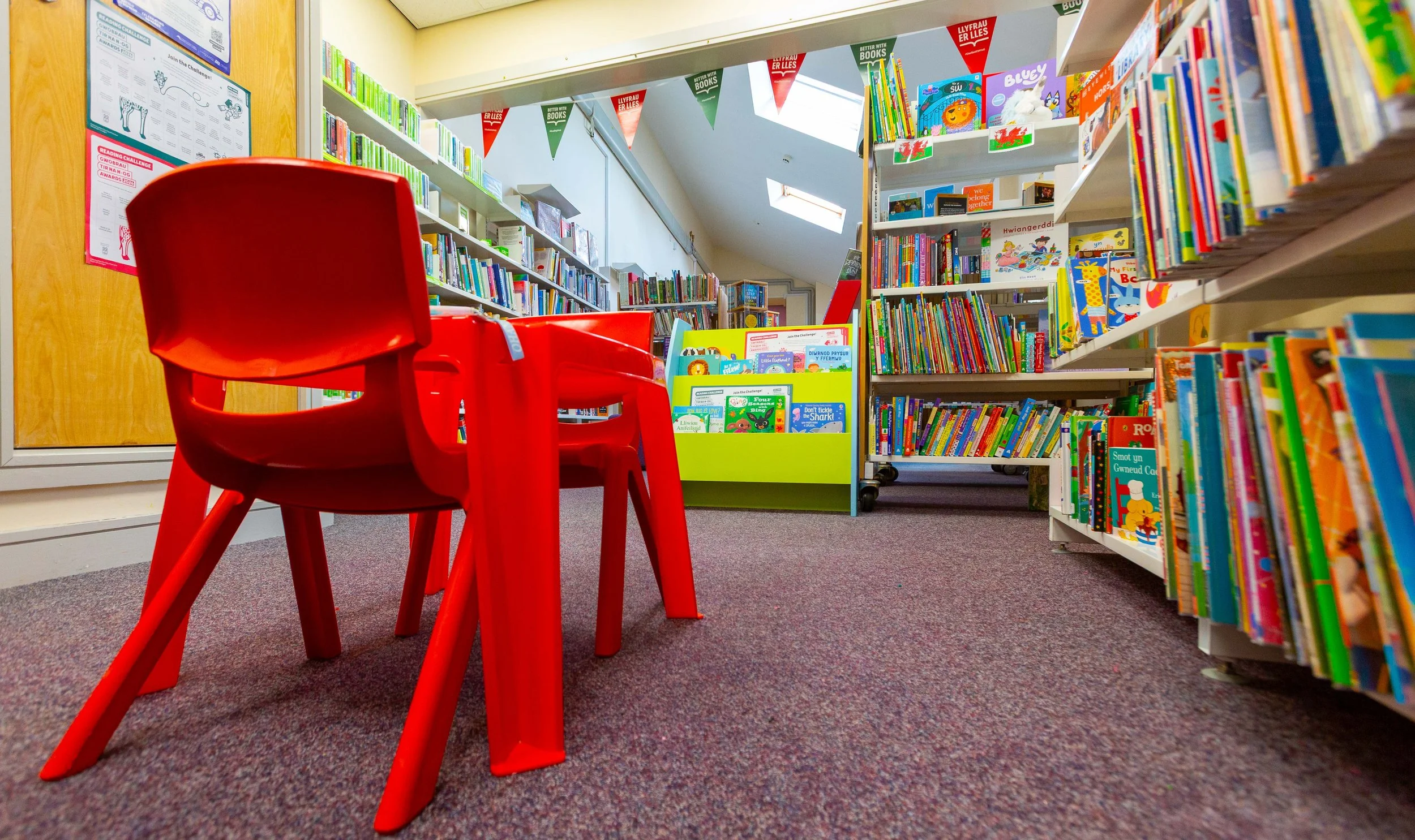 Interior of a colorful children's section in a library with bookshelf filled with children's books and a red plastic table and chairs.