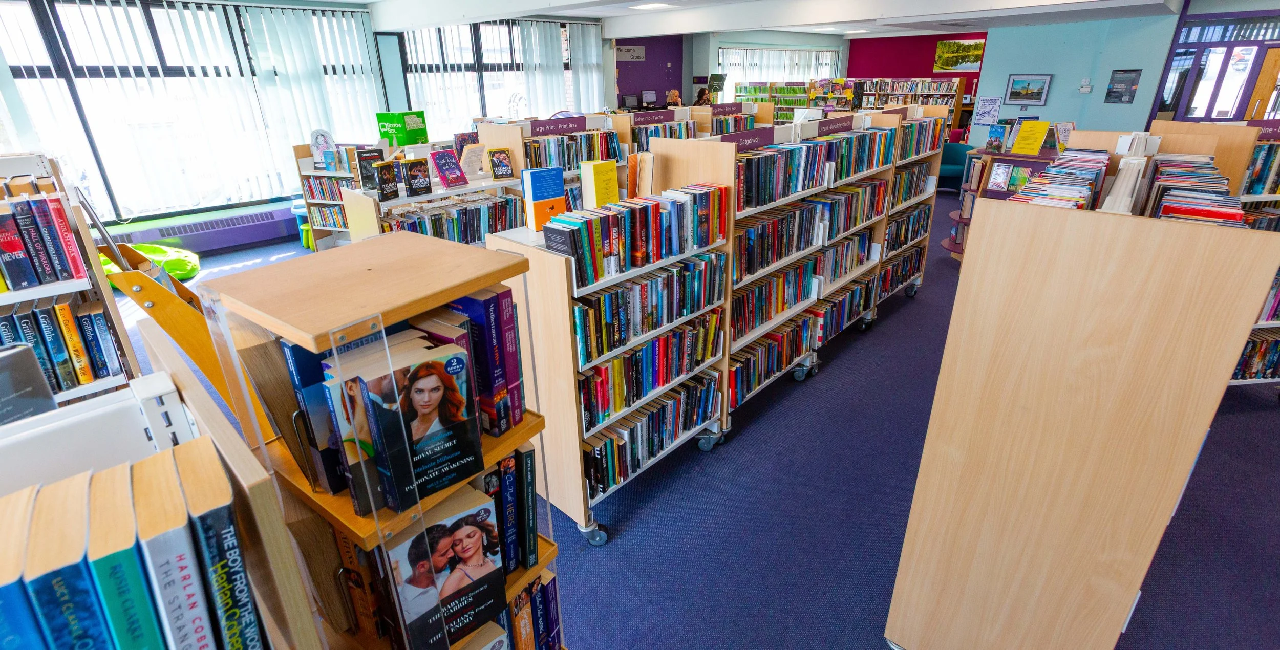 A brightly lit public library with wooden bookshelf racks filled with colorful books, large windows with vertical blinds, and a blue carpeted floor.