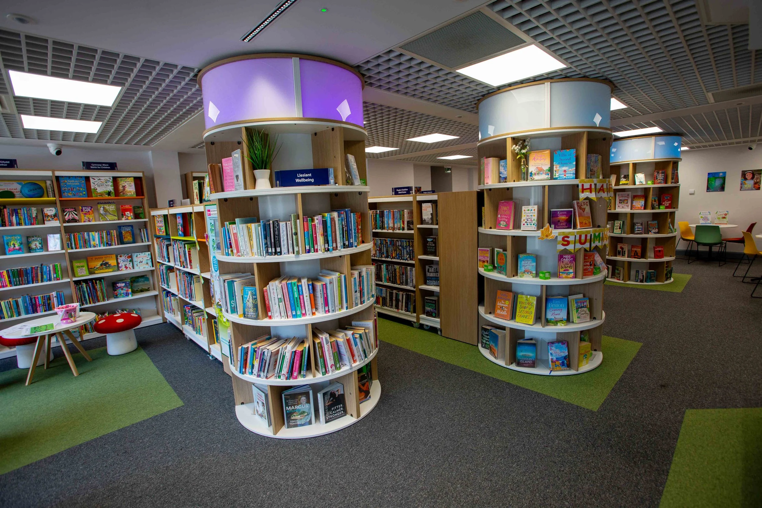 Bookshelves filled with colorful books in a library with seating areas and decorative green and gray carpeted flooring.