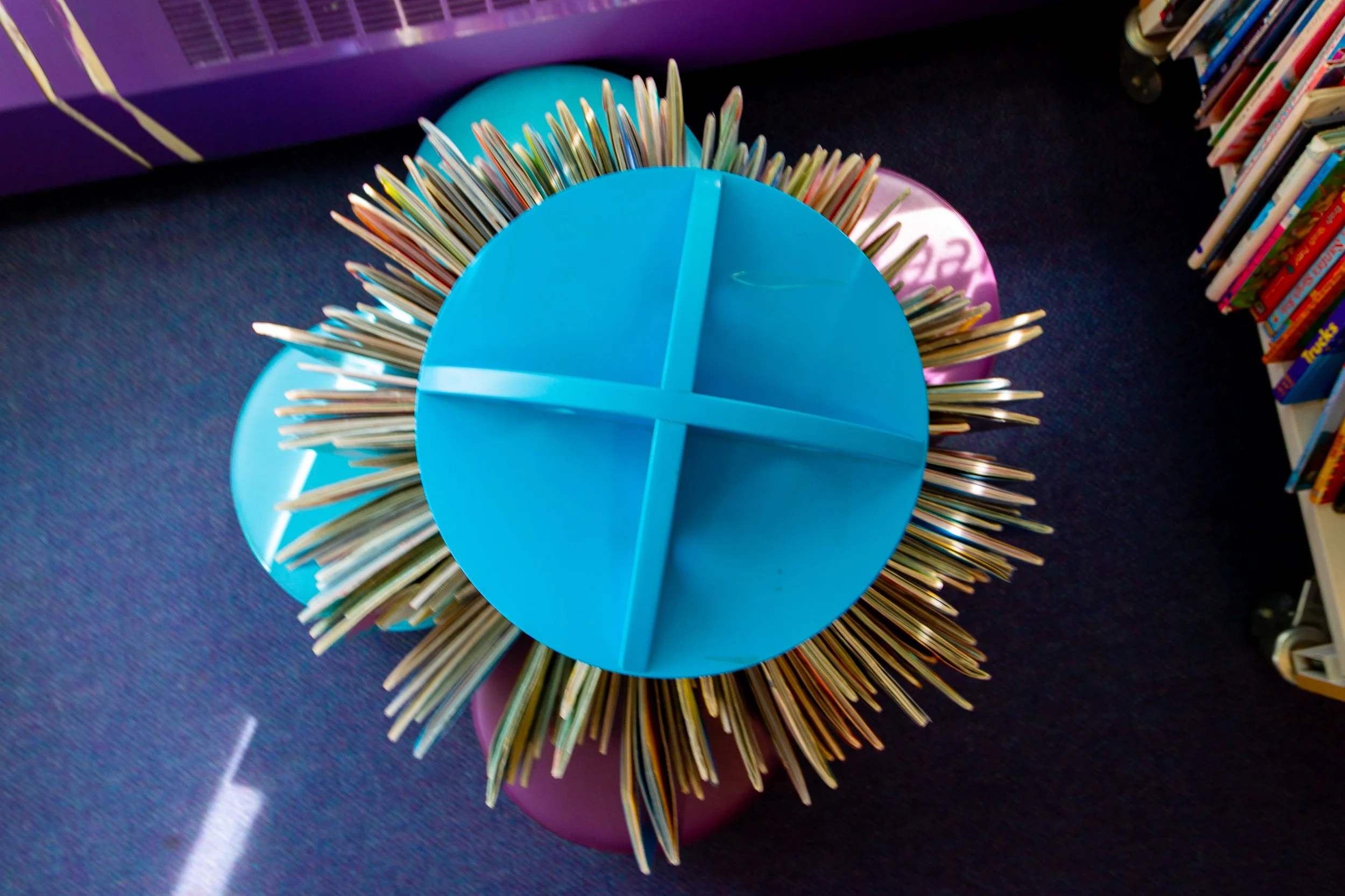A round blue table with four sections, surrounded by a pile of books, situated in a library or bookstore with shelves of books visible in the background.