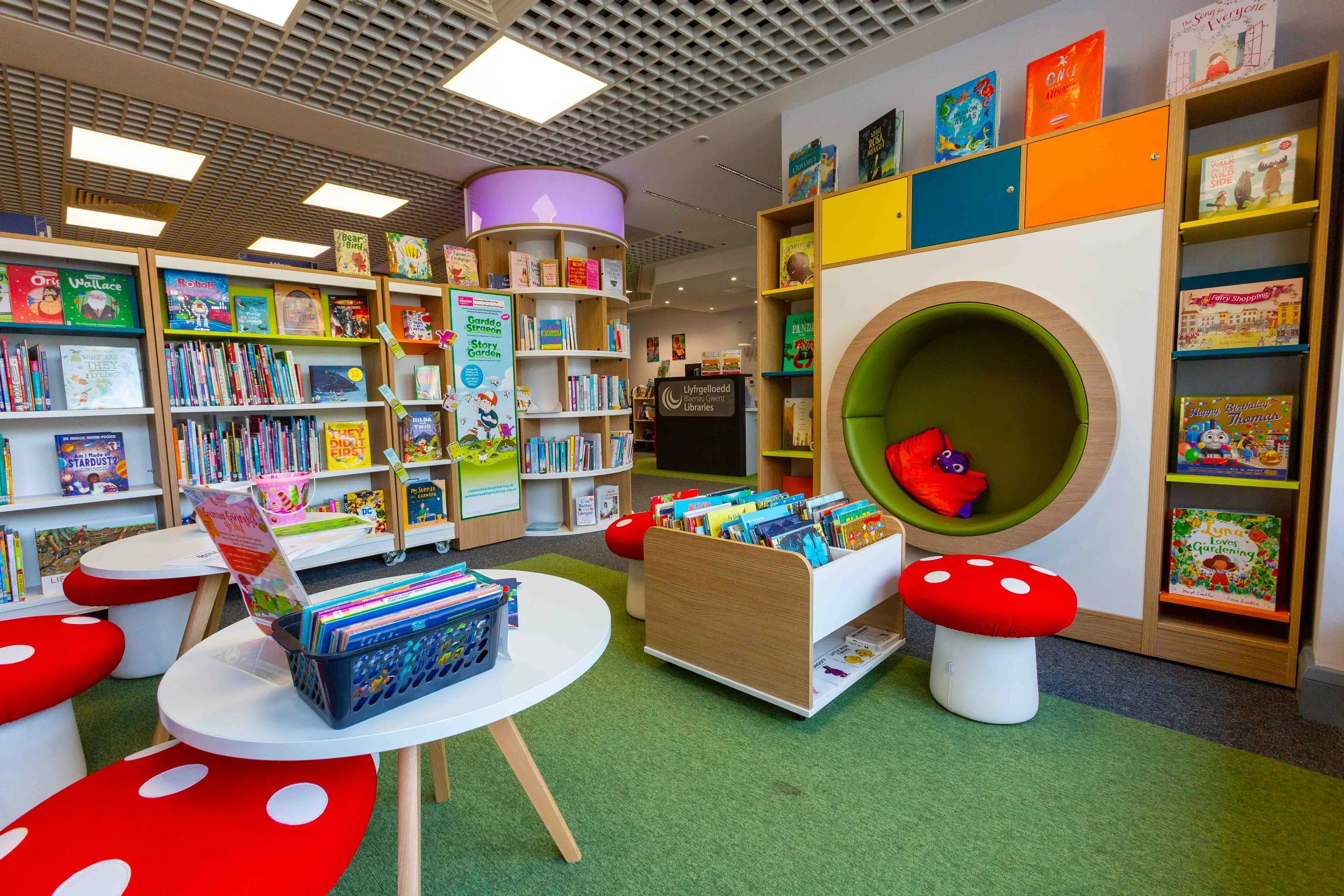 Children's section in a library featuring bookshelves filled with colorful children's books, a circular green and purple reading nook with pillows, a white table with red mushroom-shaped stools, and a cozy green carpet area.