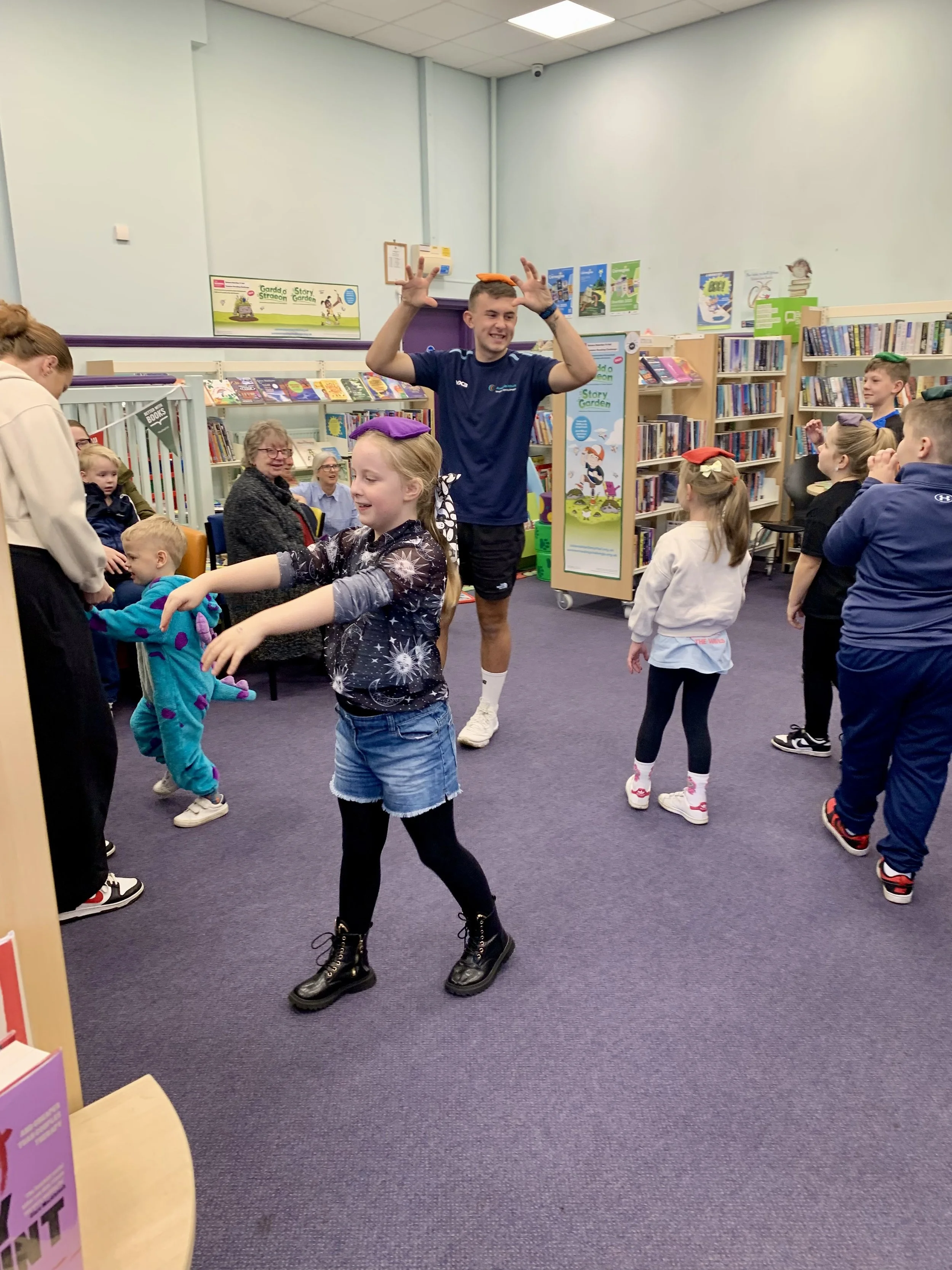 Children and adults in a library engaged in a fun activity or game, with bookshelves in the background.