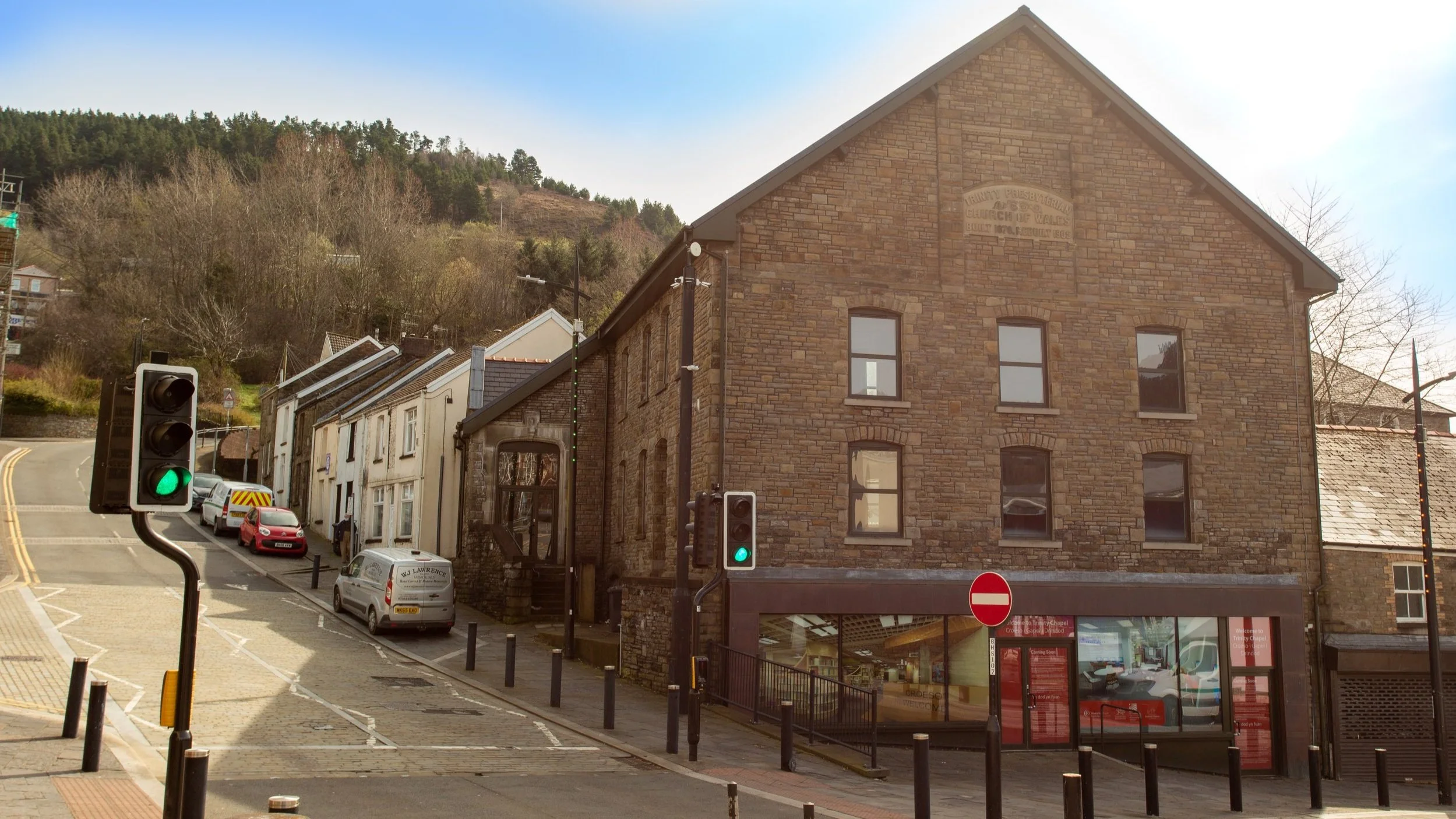A street scene with a large brick building on the right, multiple parked cars, traffic signals, and a hillside with trees in the background.