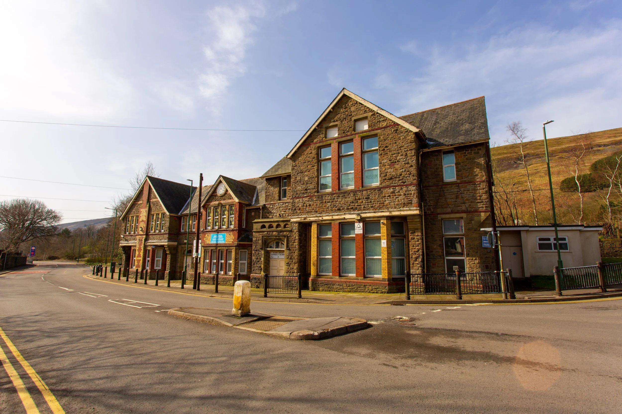 A street view showing old stone and brick buildings, with a hillside in the background, clear sky, and road markings.