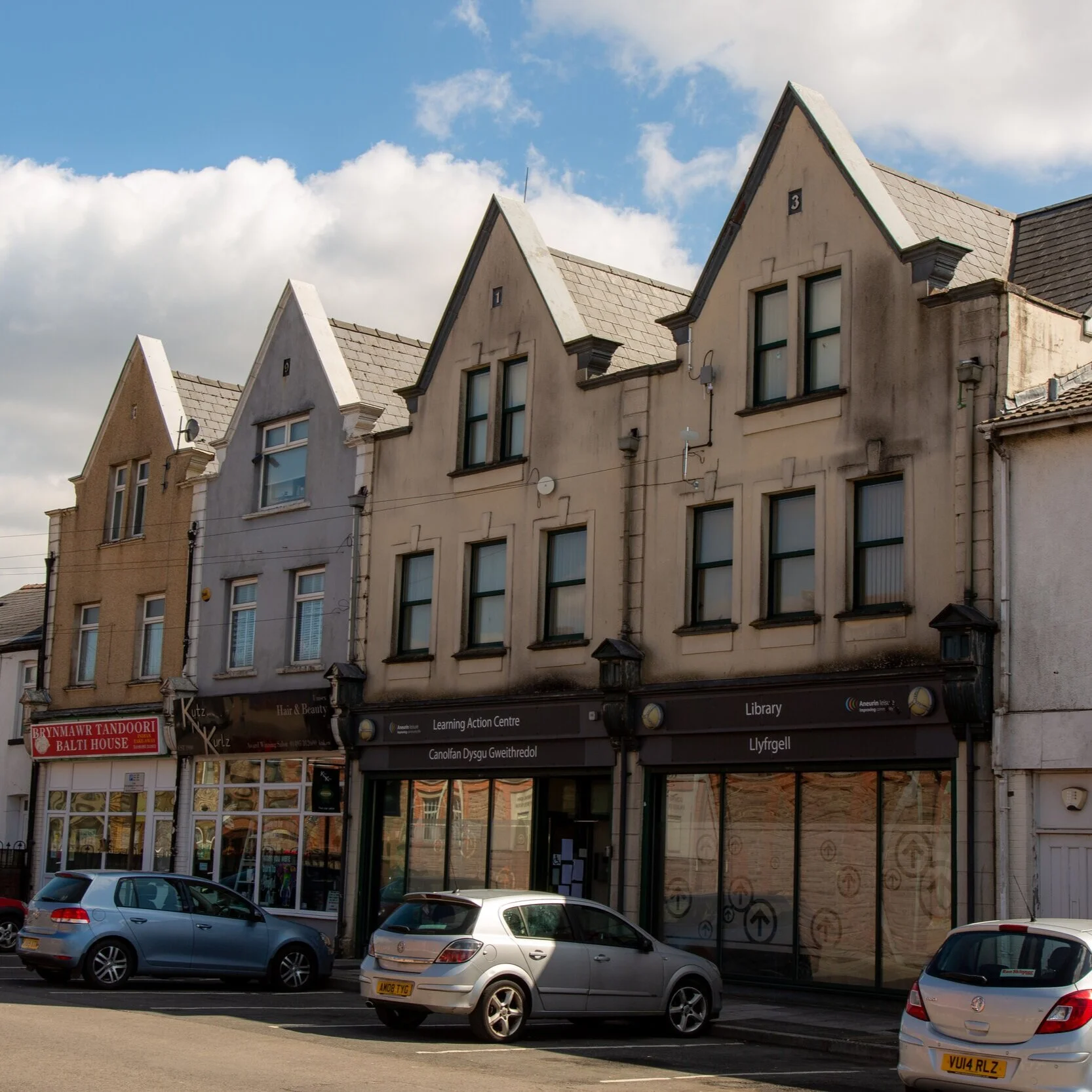 Row of old buildings with storefronts on the ground level and traditional gabled roofs, parked cars in front, under a partly cloudy sky.