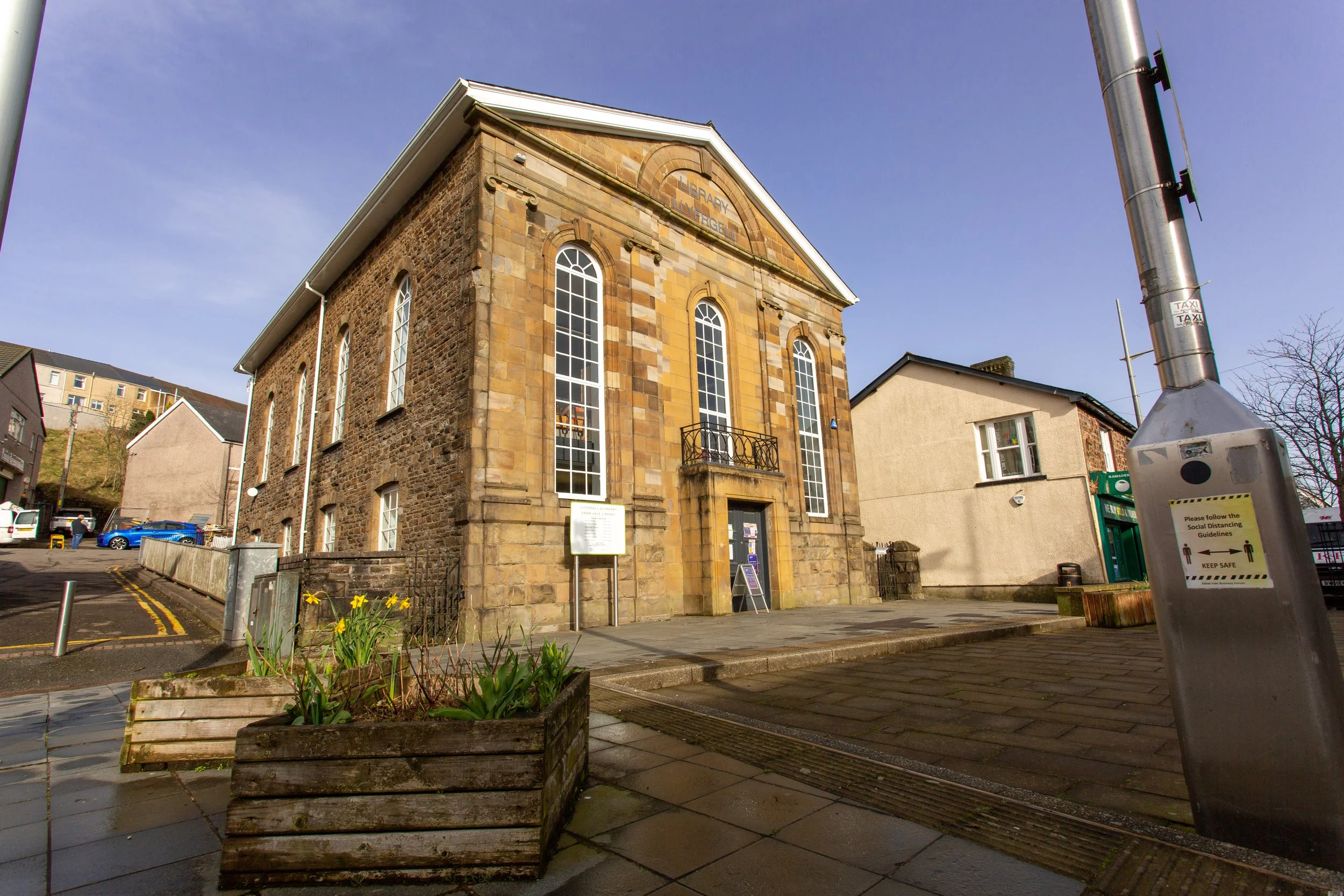 A historic stone building with tall arched windows and a small balcony on the front, labeled as a library, located on a corner with a sidewalk and some flower beds, and a sign on a stand near the entrance.