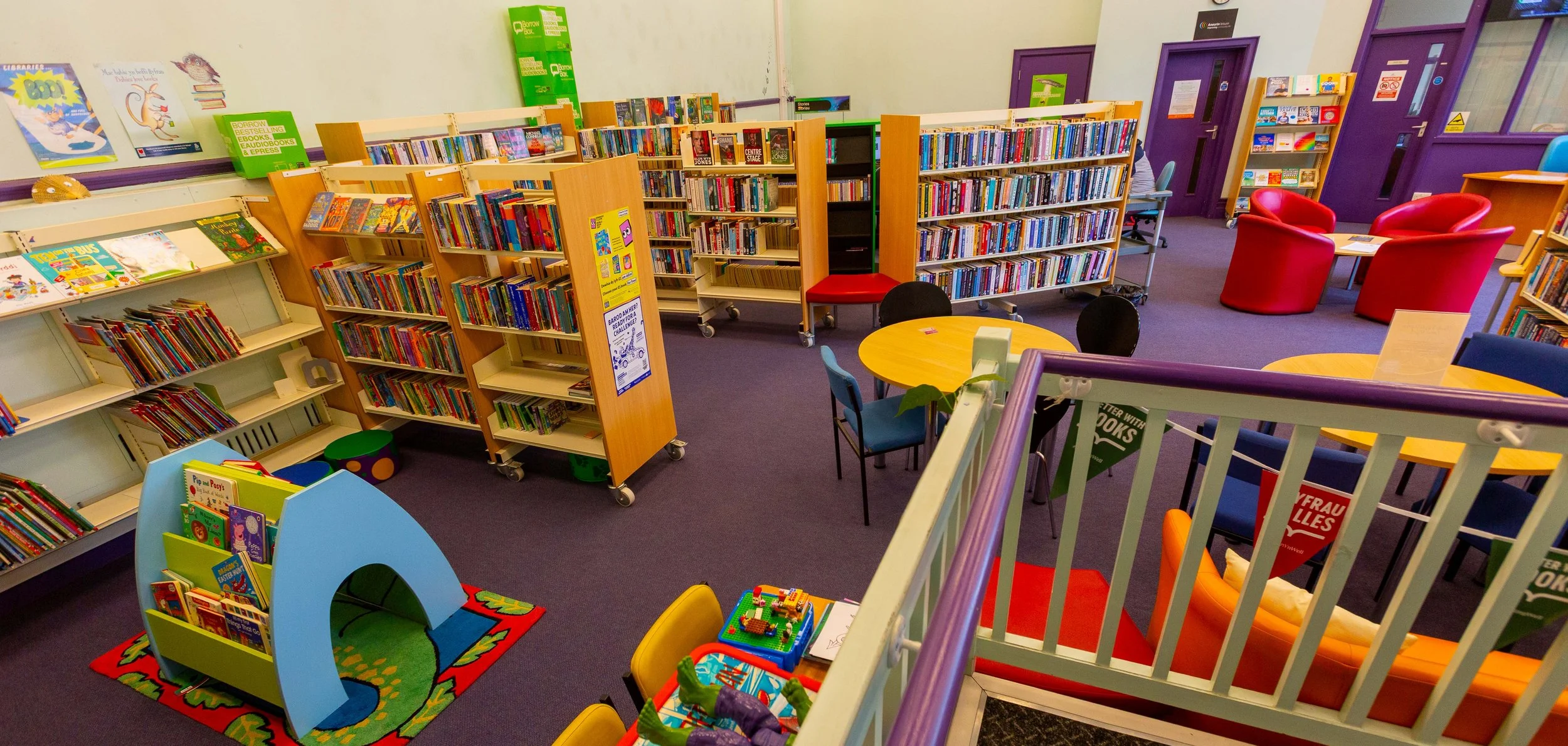Interior of a colorful children's library with bookshelves, books, and seating areas, including red, orange, and blue chairs and small tables, with purple walls and windows.