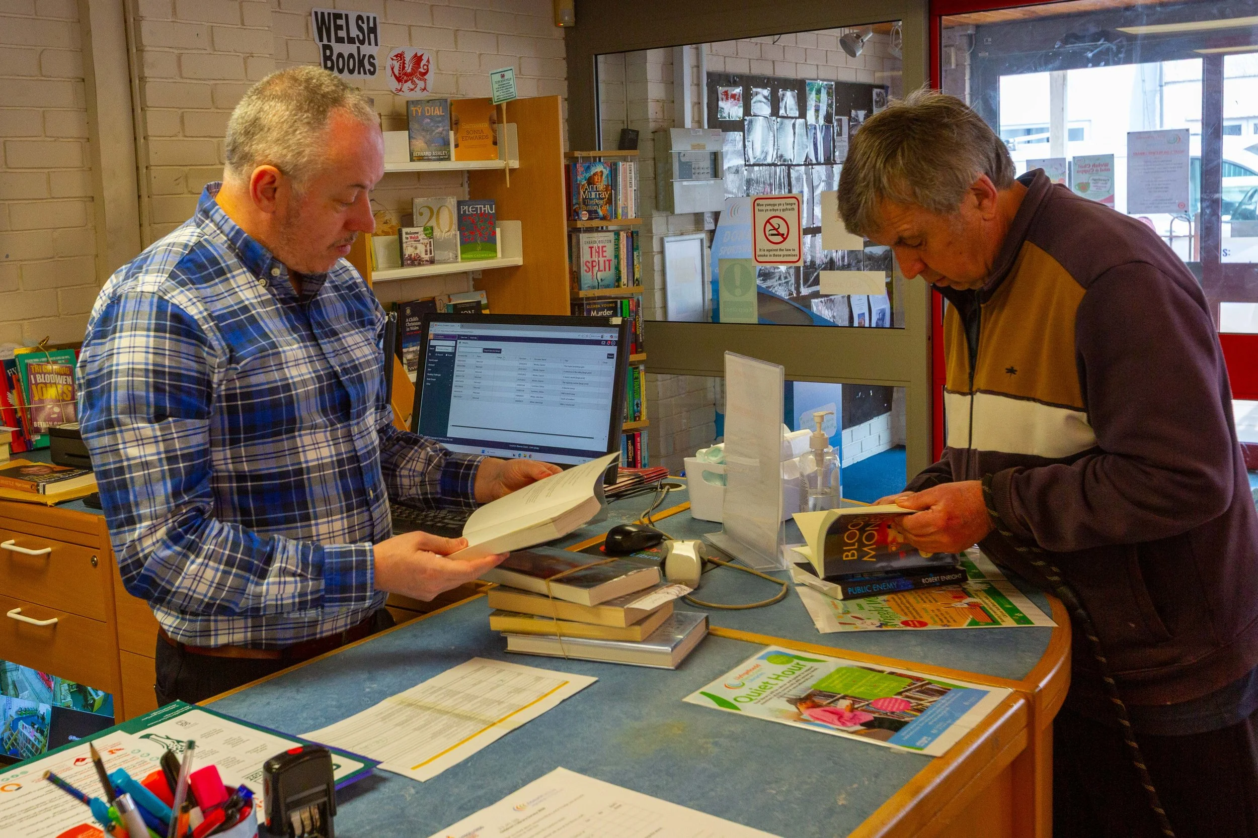 Two men standing at a counter in a bookstore, both reading books. The counter has multiple books, papers, and office supplies, with a computer monitor visible in the background. Behind the counter are bookshelves and signs, including one that says "W