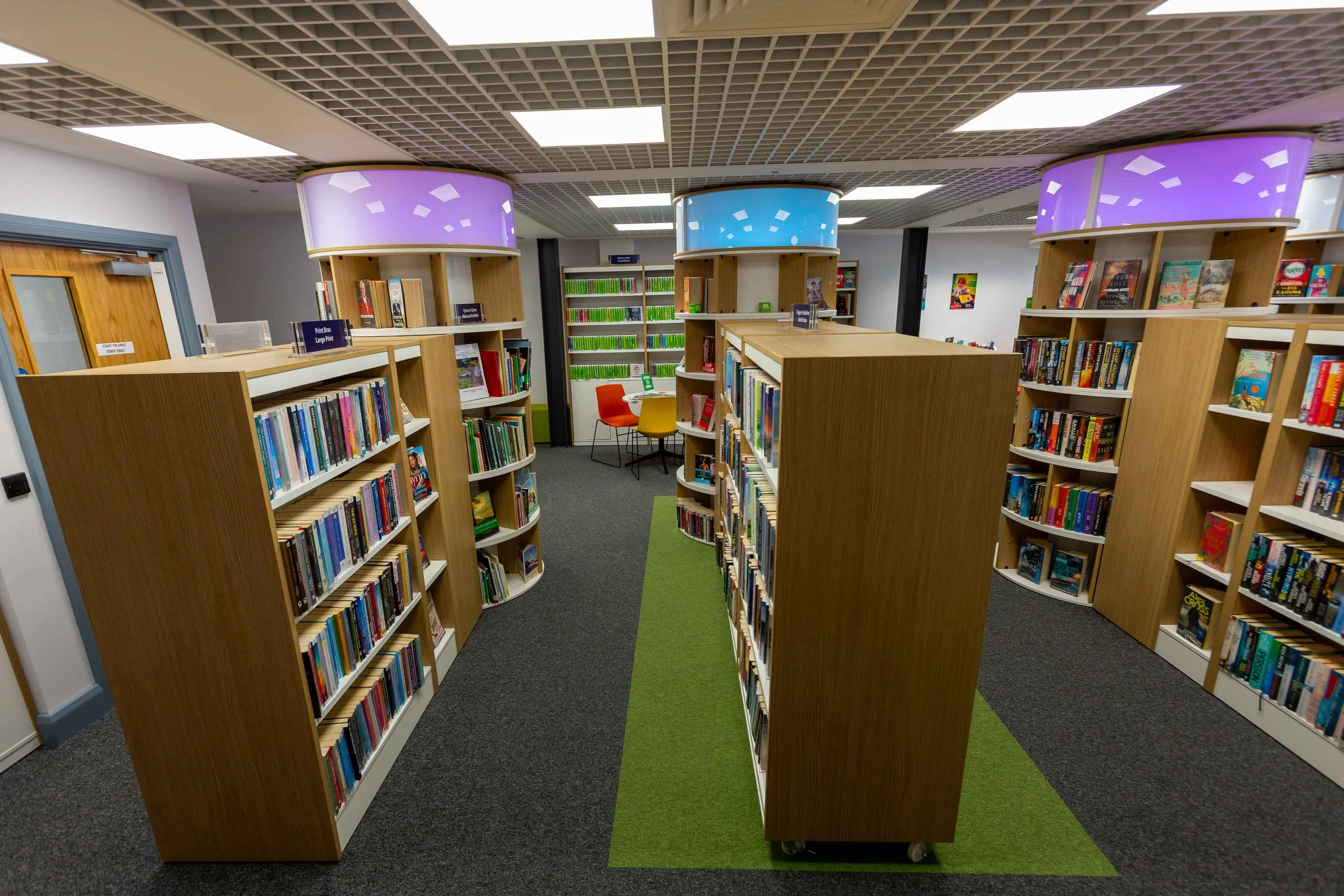 Interior of a modern library with wooden bookshelves, colorful chairs, and purple and blue illuminated ceiling fixtures.