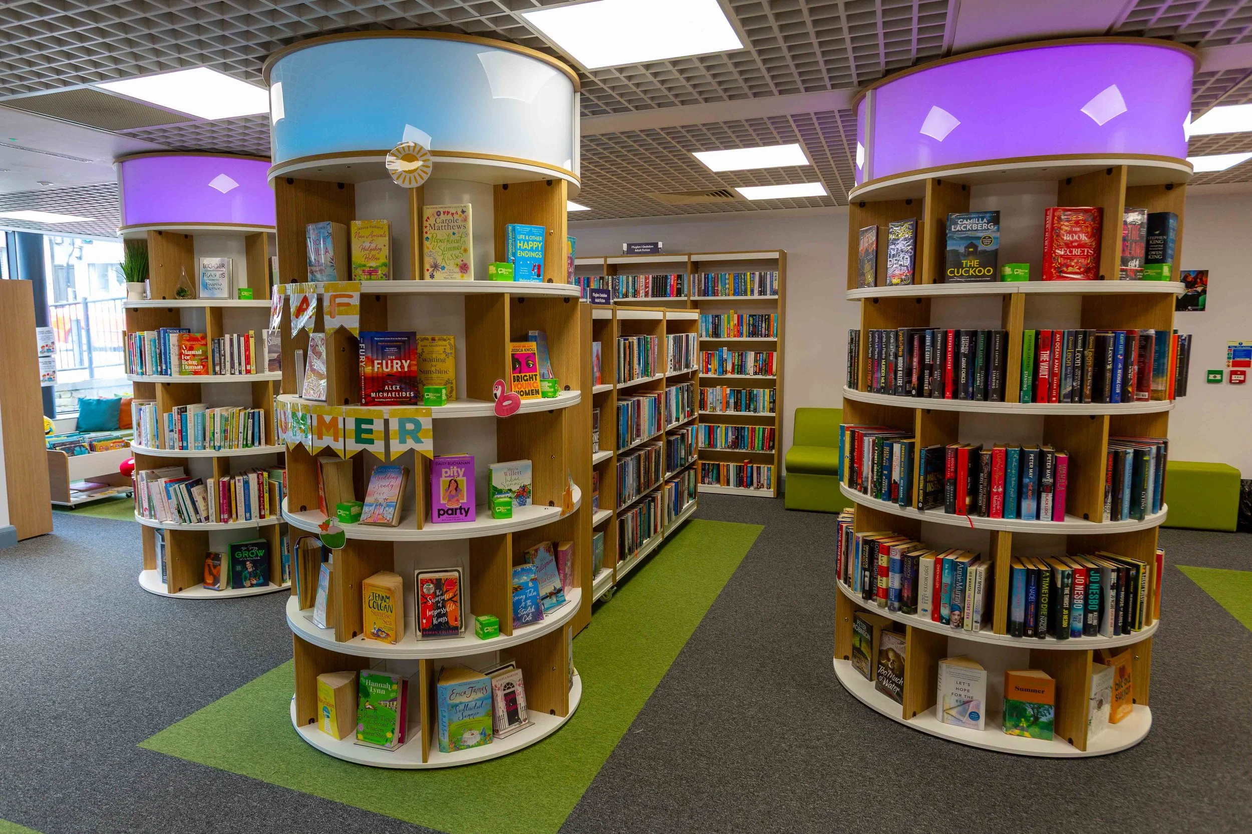 Bookshelves in a library with colorful book displays, green seating, and decorative lights on ceiling.