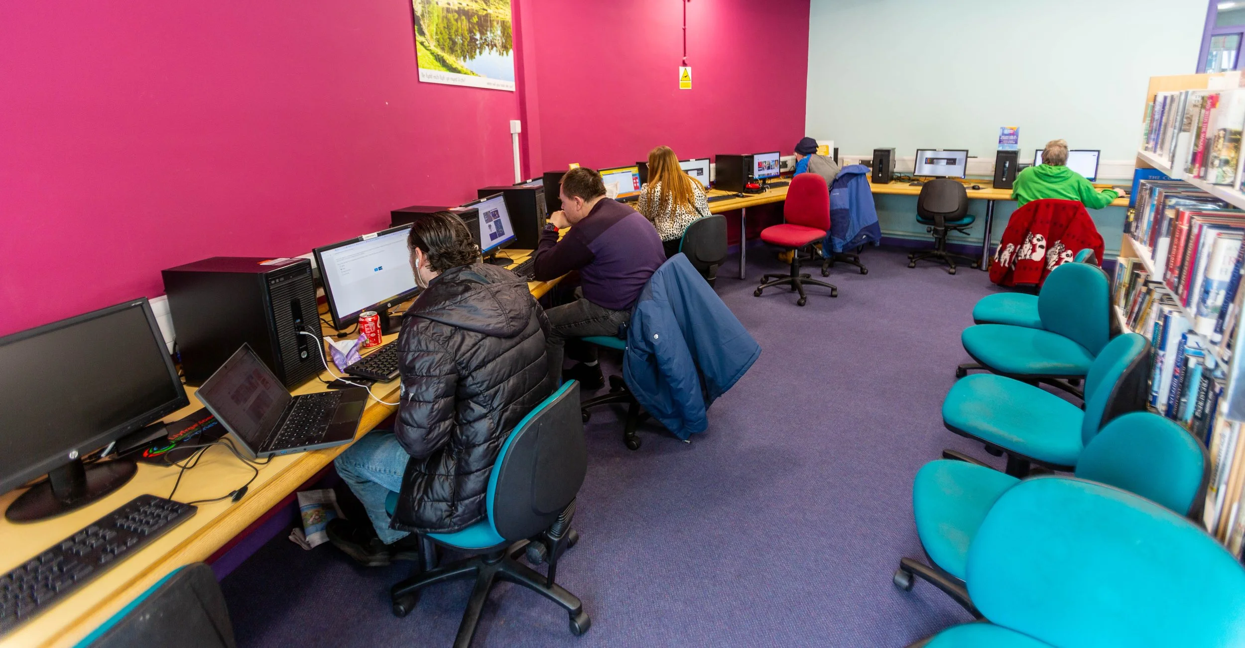 A library computer room with multiple people working on desktop computers along the purple wall. There are bookshelves on the right, and the chairs in the foreground are unoccupied.