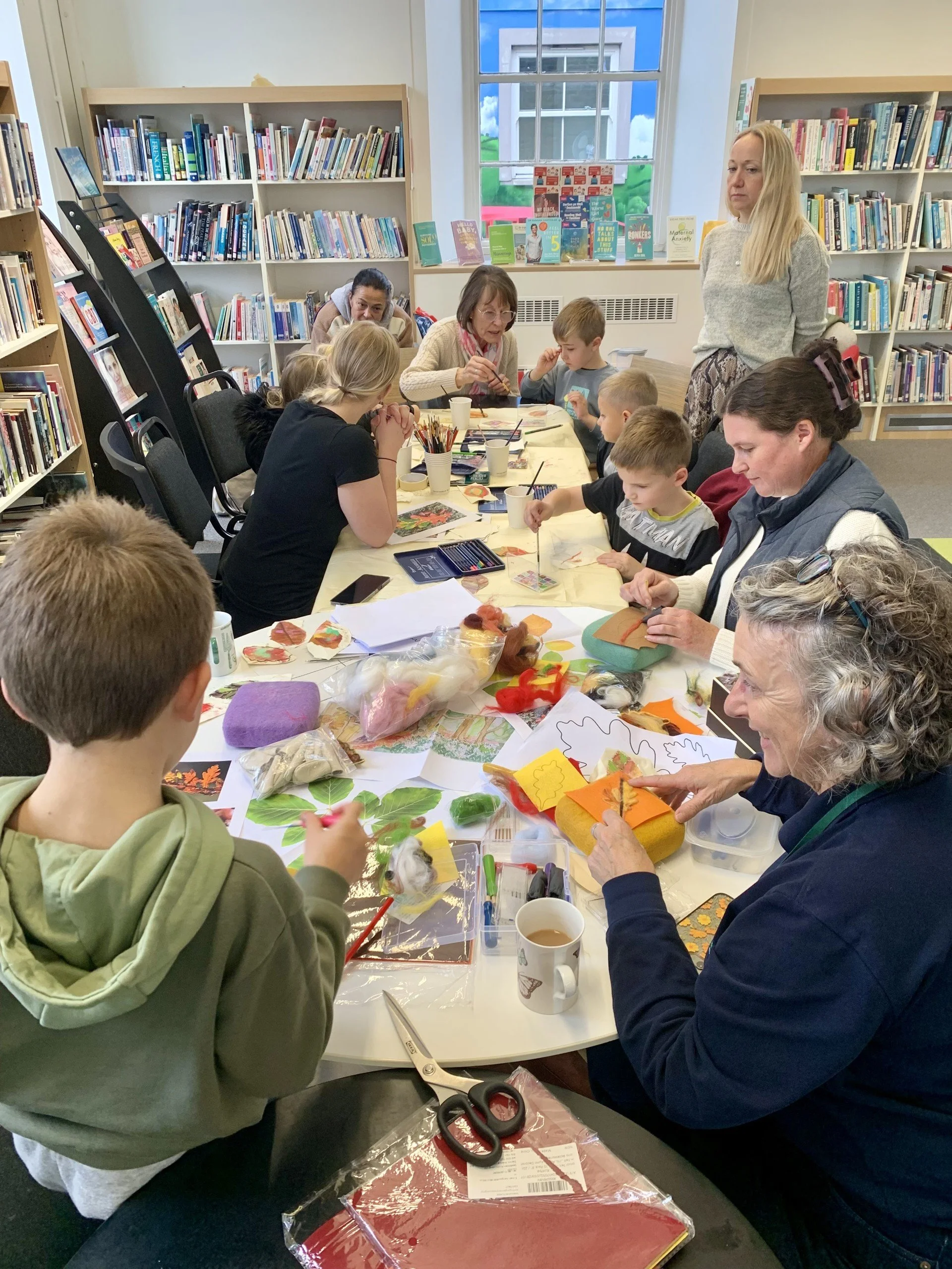 People participating in a craft workshop around a table in a library, with children and adults working on arts and crafts projects, surrounded by bookshelves and natural light from a window.