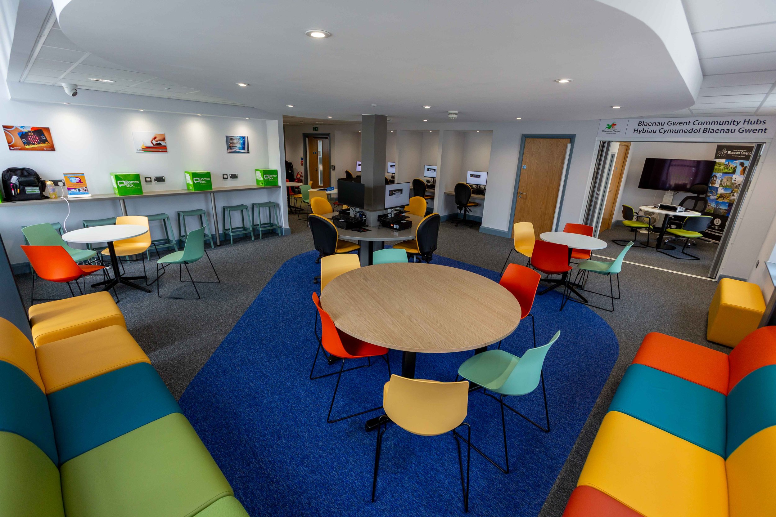 A colorful community hub with round tables, chairs, seating area, and computer stations.