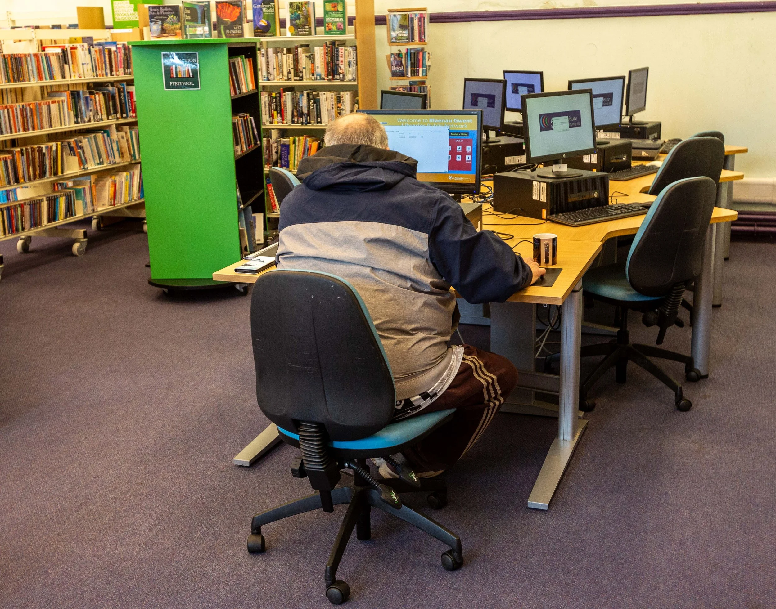 A person sitting at a computer station in a library with bookshelves and multiple desktop computers.
