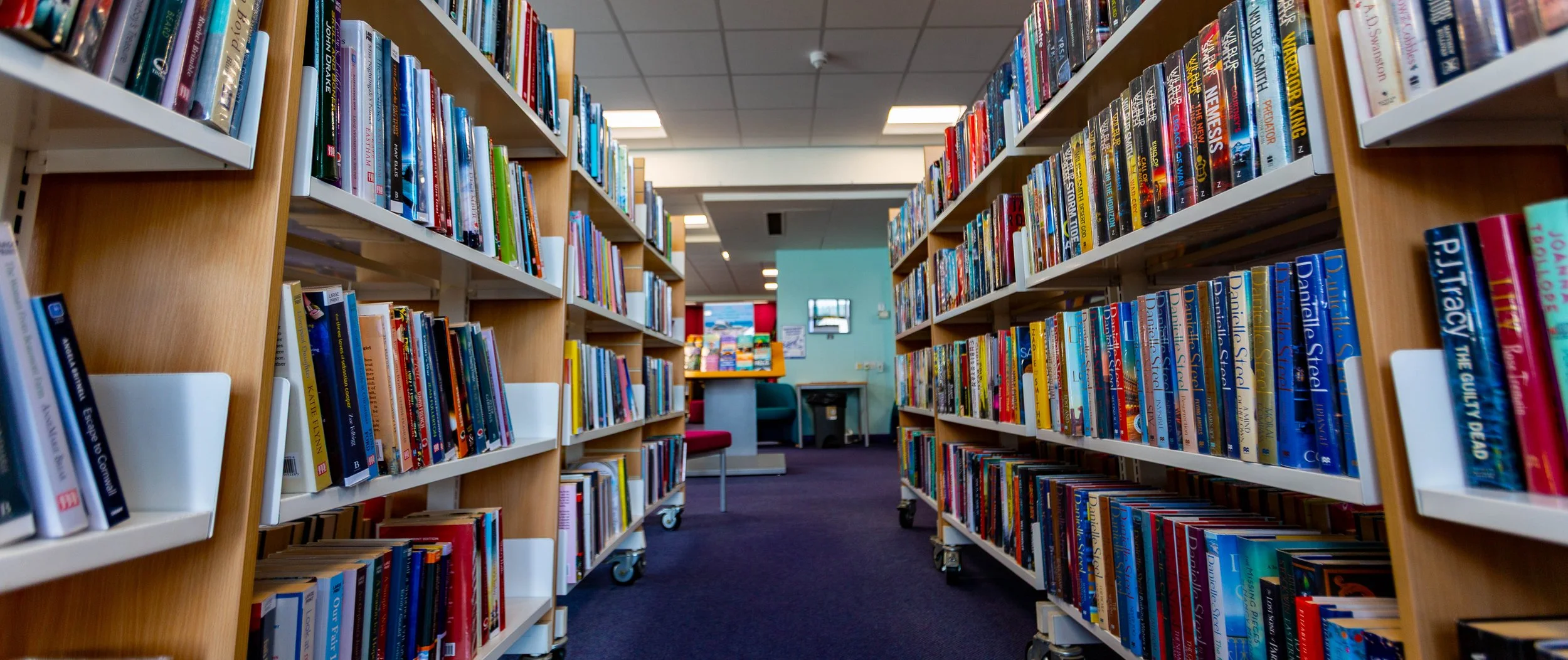 Bookshelves filled with books in a library aisle, with a reading table and chairs visible in the background.