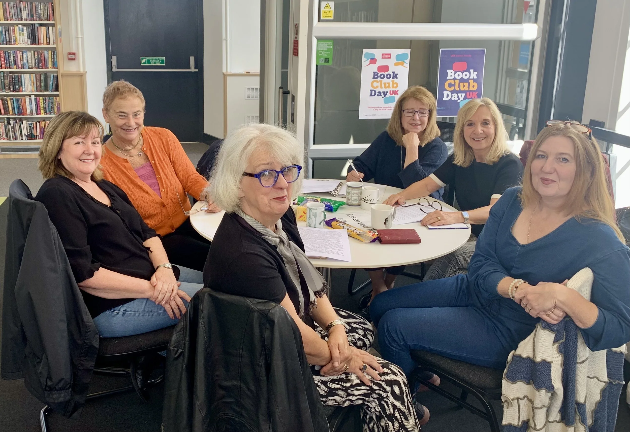 A group of seven women sitting around a table in a library or community center during Book Club Day UK. They are smiling and have books, papers, mugs, and snacks on the table. Posters on the window behind them display the event name.