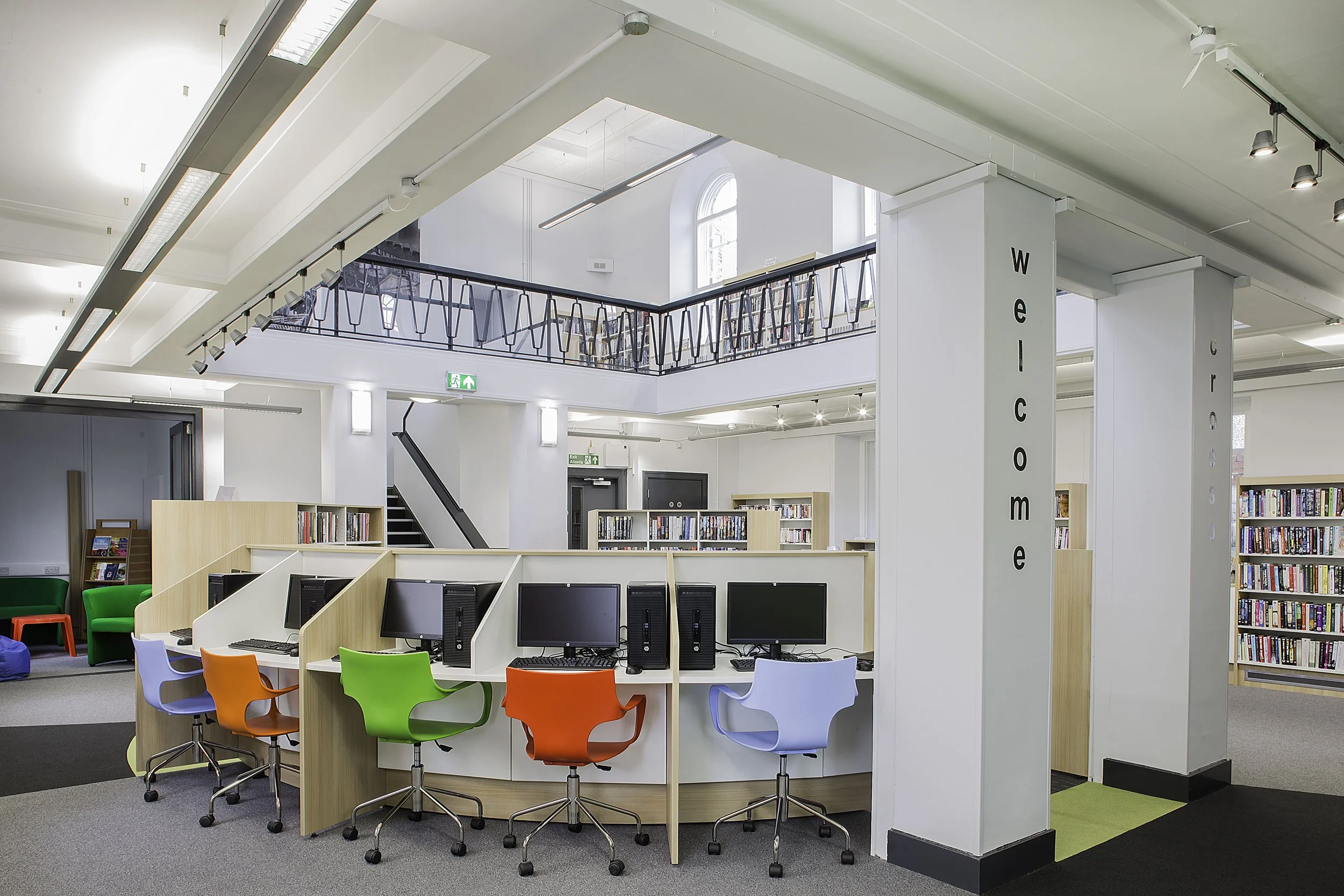 Interior of a modern library with a computer station, colorful chairs, bookshelves, and a mezzanine level.