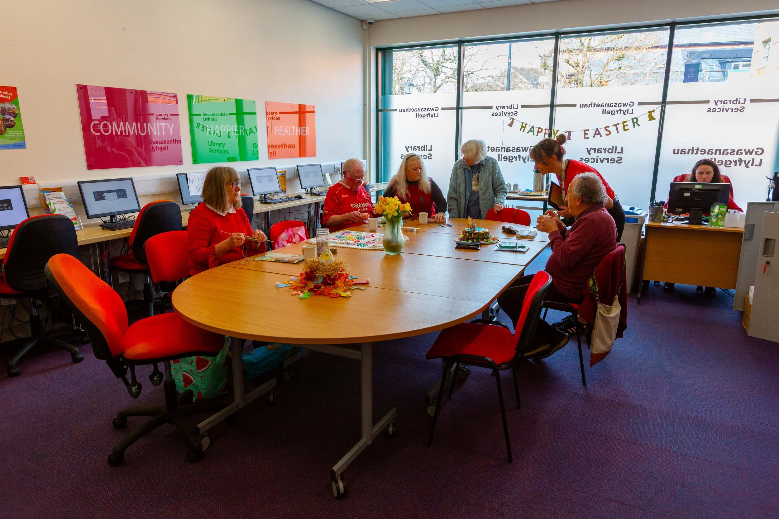 People gathered around a large table in a room decorated for Easter, with some working on crafts and a woman assisting an elderly man, in a community center or library setting with colorful posters and computer stations.