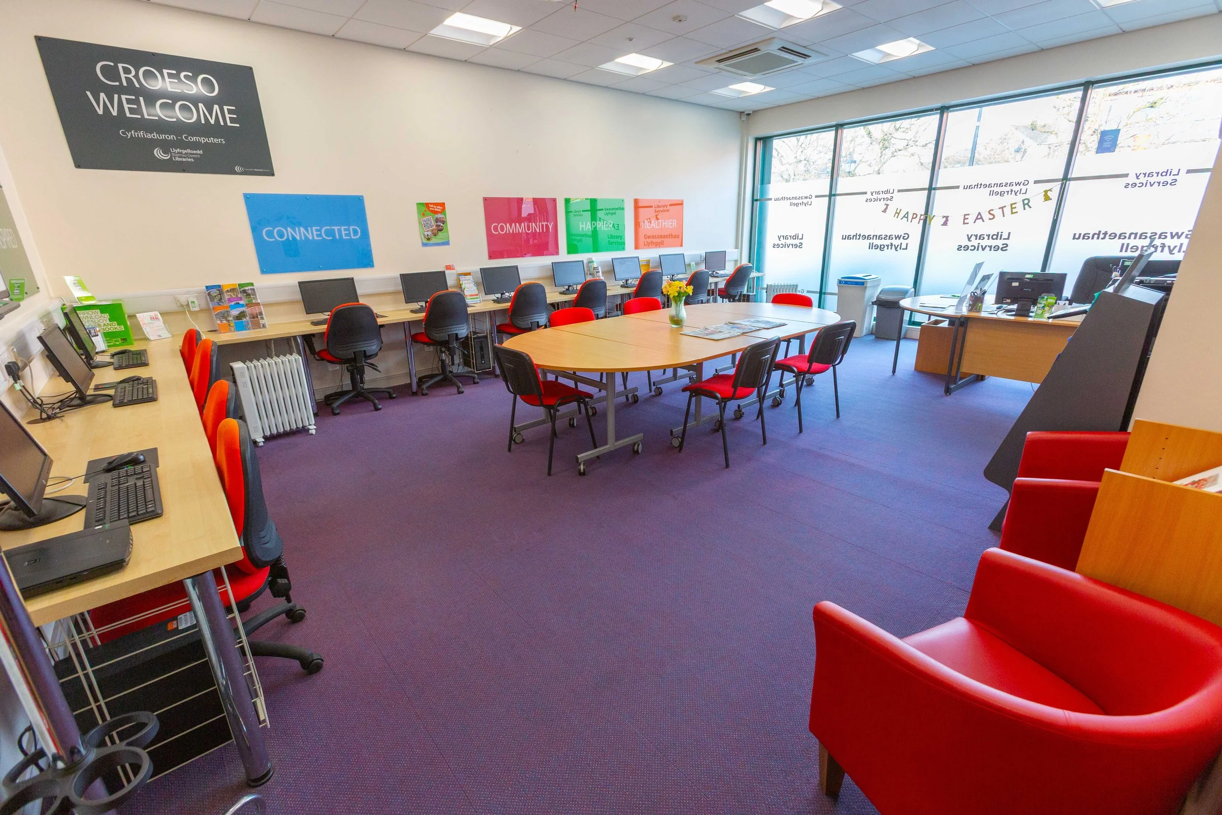 Bright library or community center room with computers along the wall, tables with chairs in the center, and large windows with 'Happy Easter' decorations on the glass.
