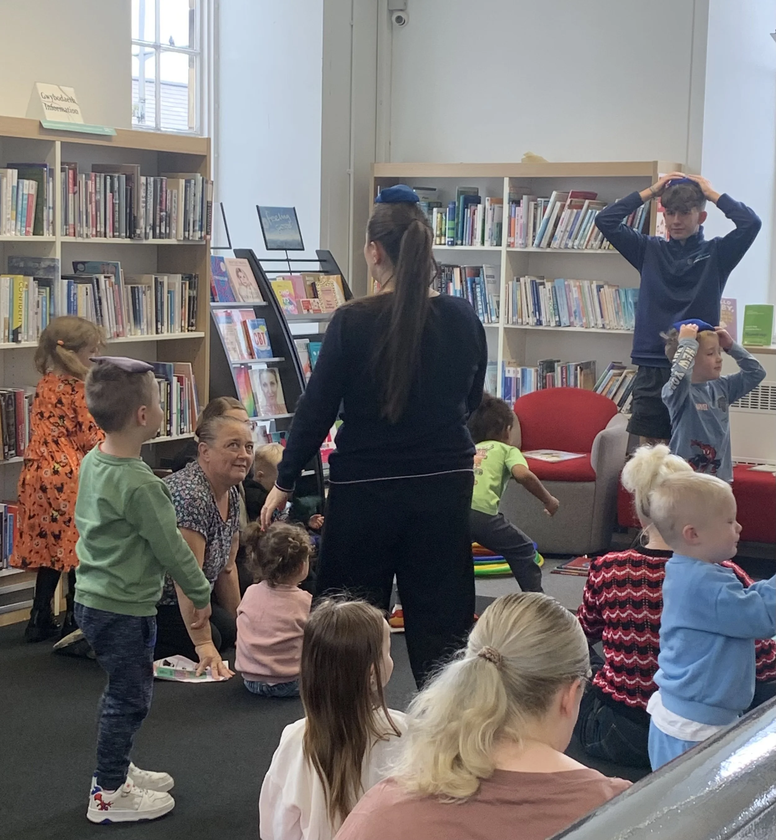 Children and adults gathered in a library for a storytelling or reading event, with bookshelves and a display of books in the background.