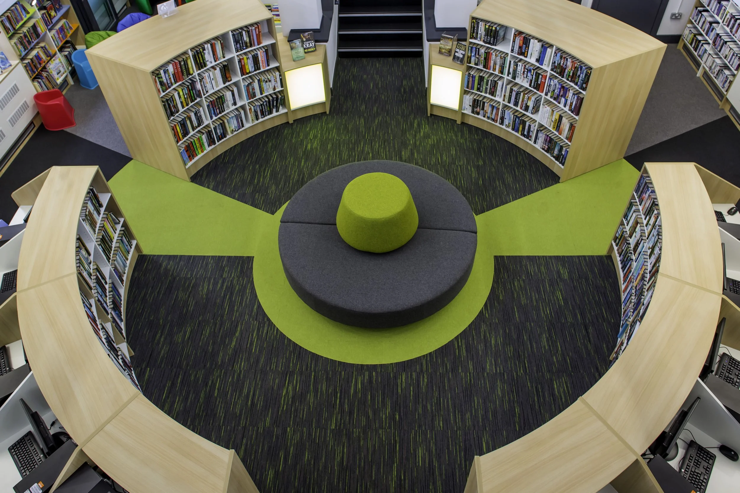 A modern library with a circular seating area in the center, surrounded by bookshelves filled with books. The seating includes a large, round, dark gray cushion with a smaller green cushion on top, on a green and gray carpet. There are computer stati