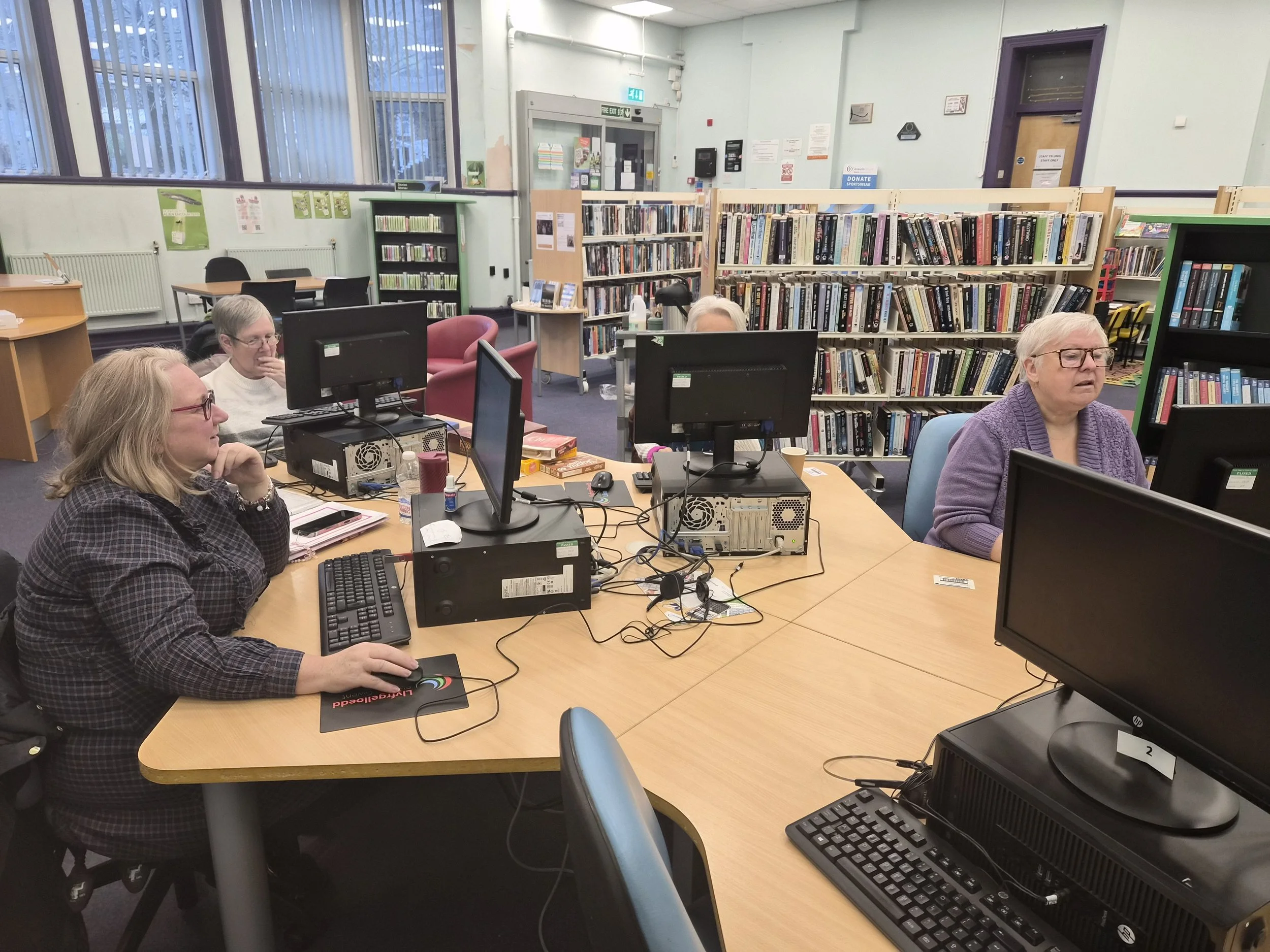 Four women working on computers in a library or computer room, with bookshelves in the background and large windows providing natural light.