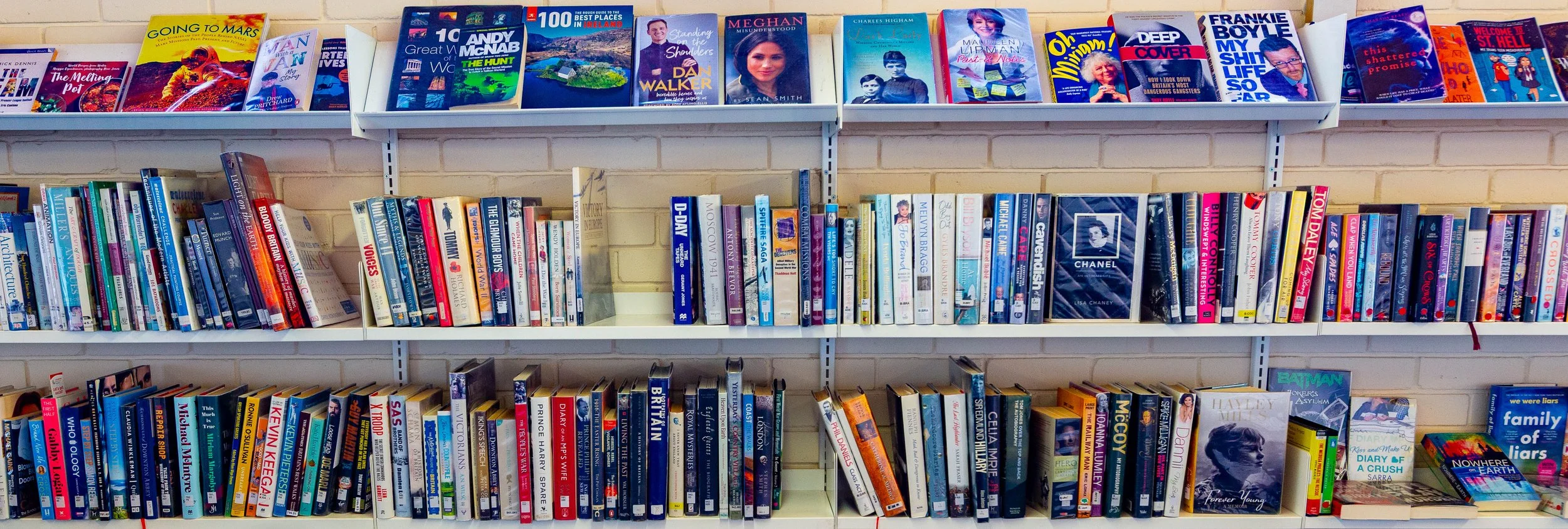 Bookshelves filled with a variety of colorful paperback books in a library. The top shelf has fiction and non-fiction titles, while the middle and bottom shelves contain a mix of novels and graphic novels.
