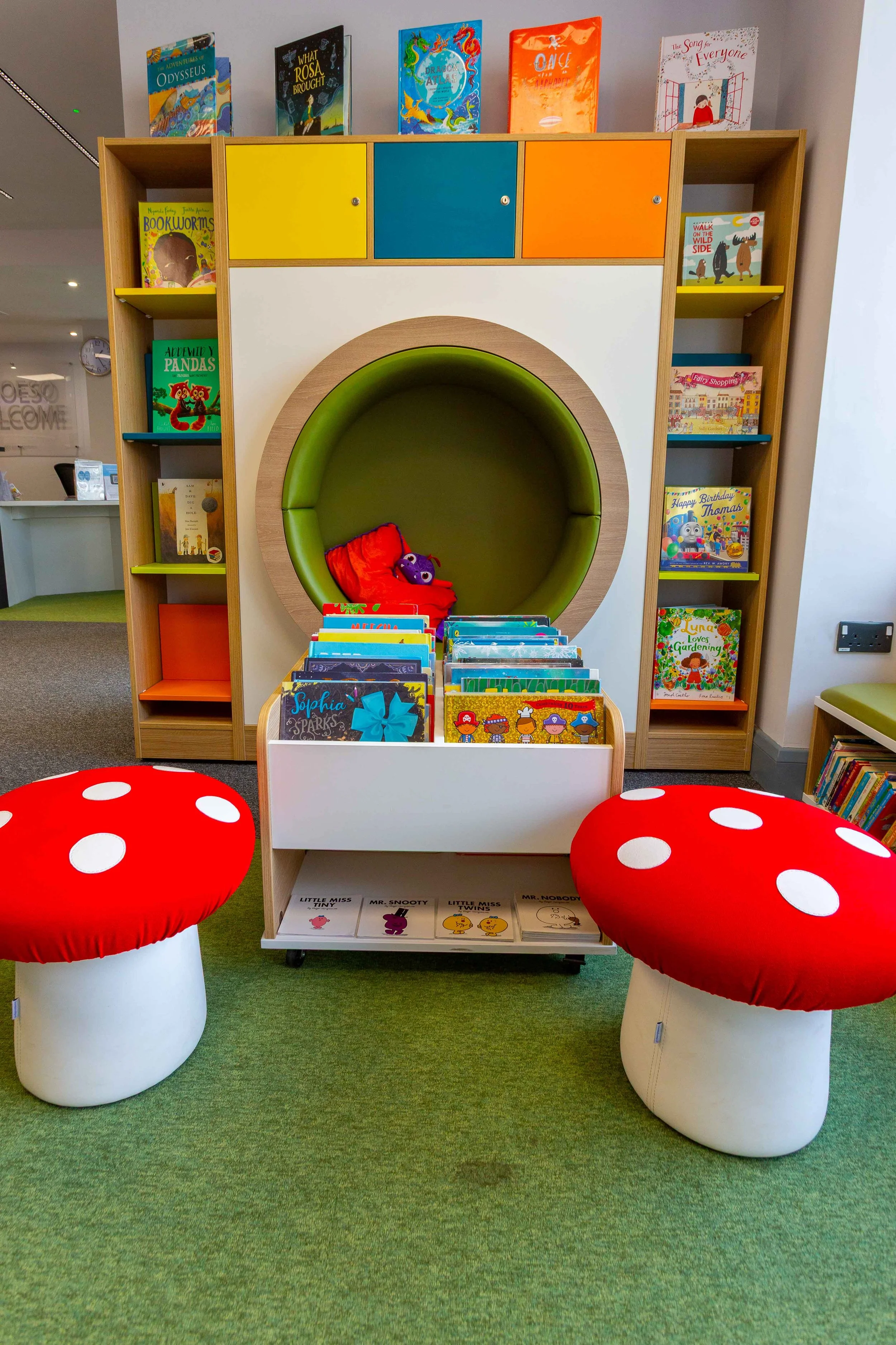 Children's library reading nook with two red-and-white mushroom-shaped chairs in front of a book display. Behind the display, there is a large circular green cushioned seat with a red pillow and purple plush toy. The background features a bookshelf with colorful books and a row of children’s books displayed on top.