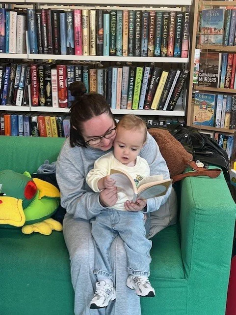 A woman with glasses and a bun is sitting on a green couch, reading a book to a young child. The child is looking at the book with interest. Behind them are shelves full of books in a library or bookstore. There is a plush frog toy next to them on th