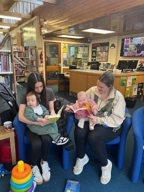 Two women with two babies sitting on bright blue chairs in a library, reading books together.