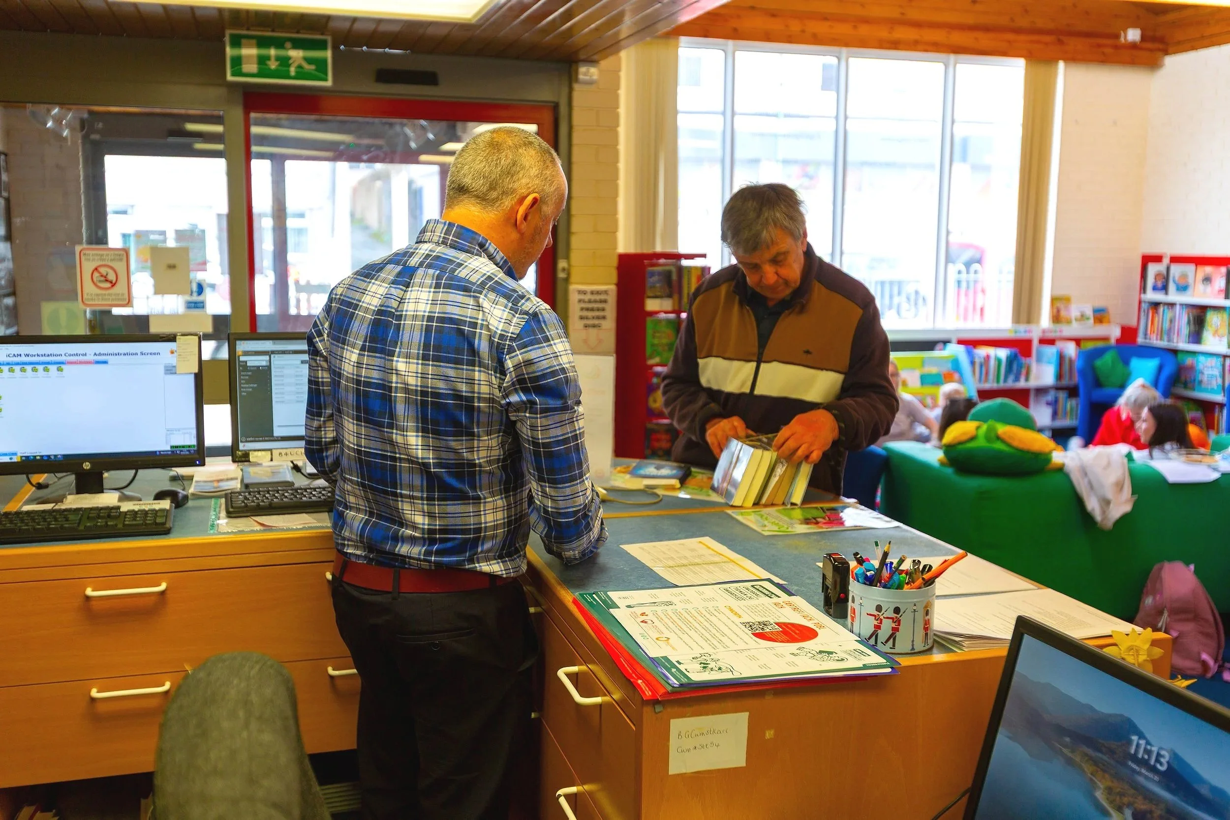 Two men at a reception desk in a library or community center. One is facing away from the camera and the other is looking at a book. In the background, there are children reading and a colorful bookshelf.