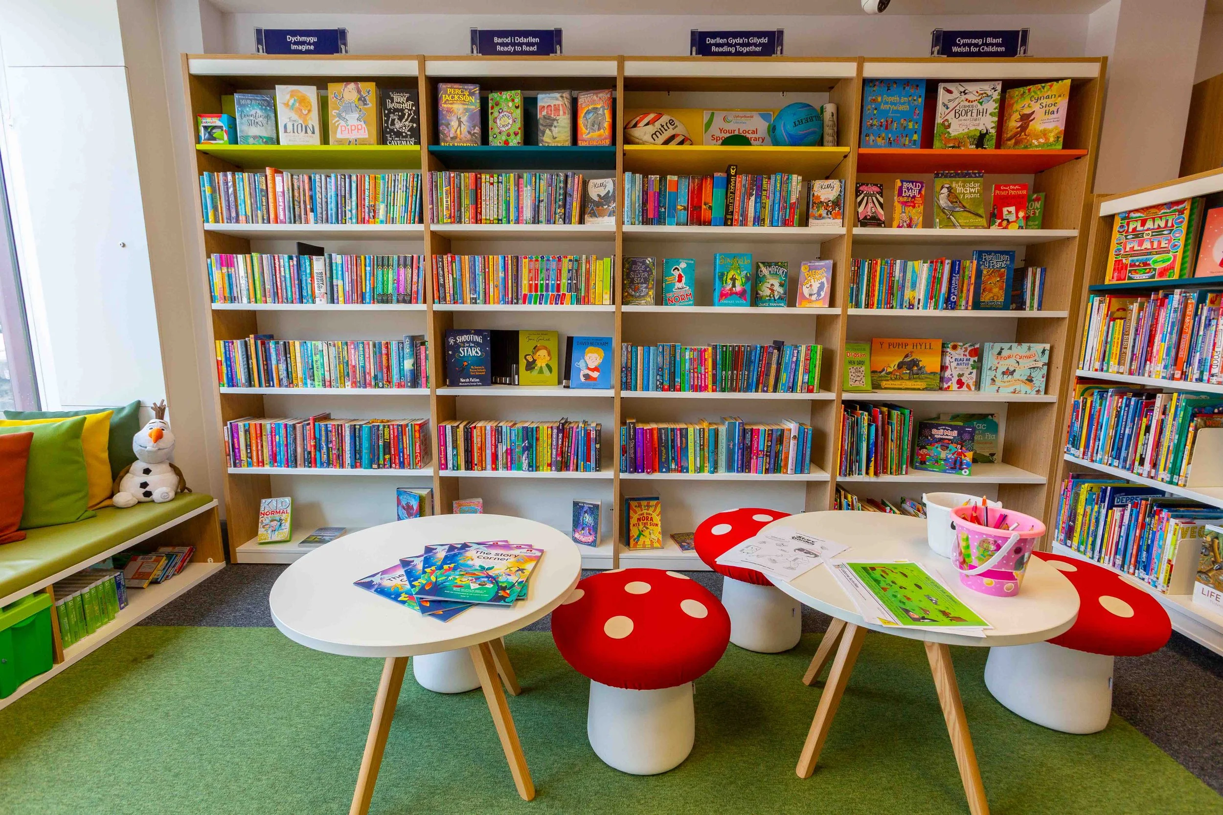 Children's library corner with colorful bookshelves filled with children's books, seating area with small tables and mushroom-shaped stools, and a plush Olaf toy on a green bench with orange and green cushions.