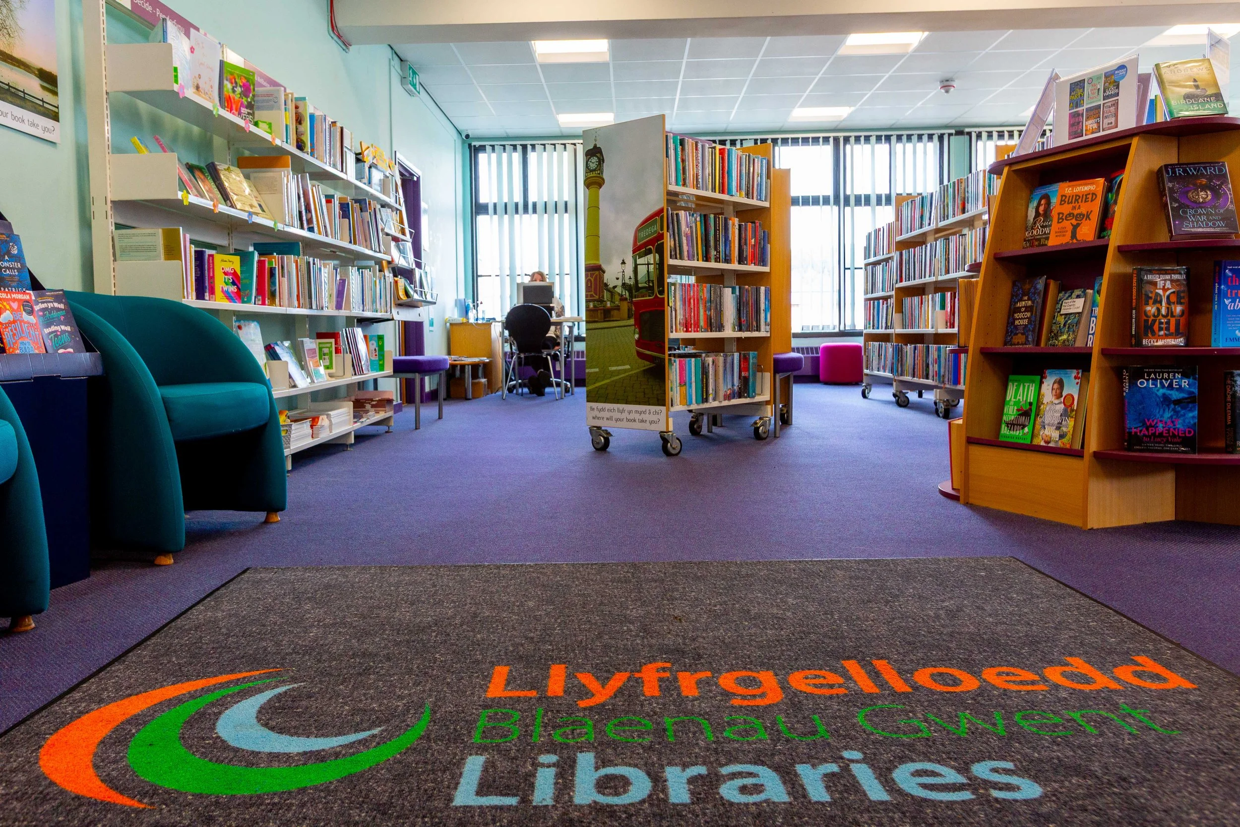 Indoor view of a library with shelves of books, colorful chairs, and a paper sign on a cart. The carpet features the Llyfrgelload Blaenau Gwent Libraries logo in orange, green, and blue.