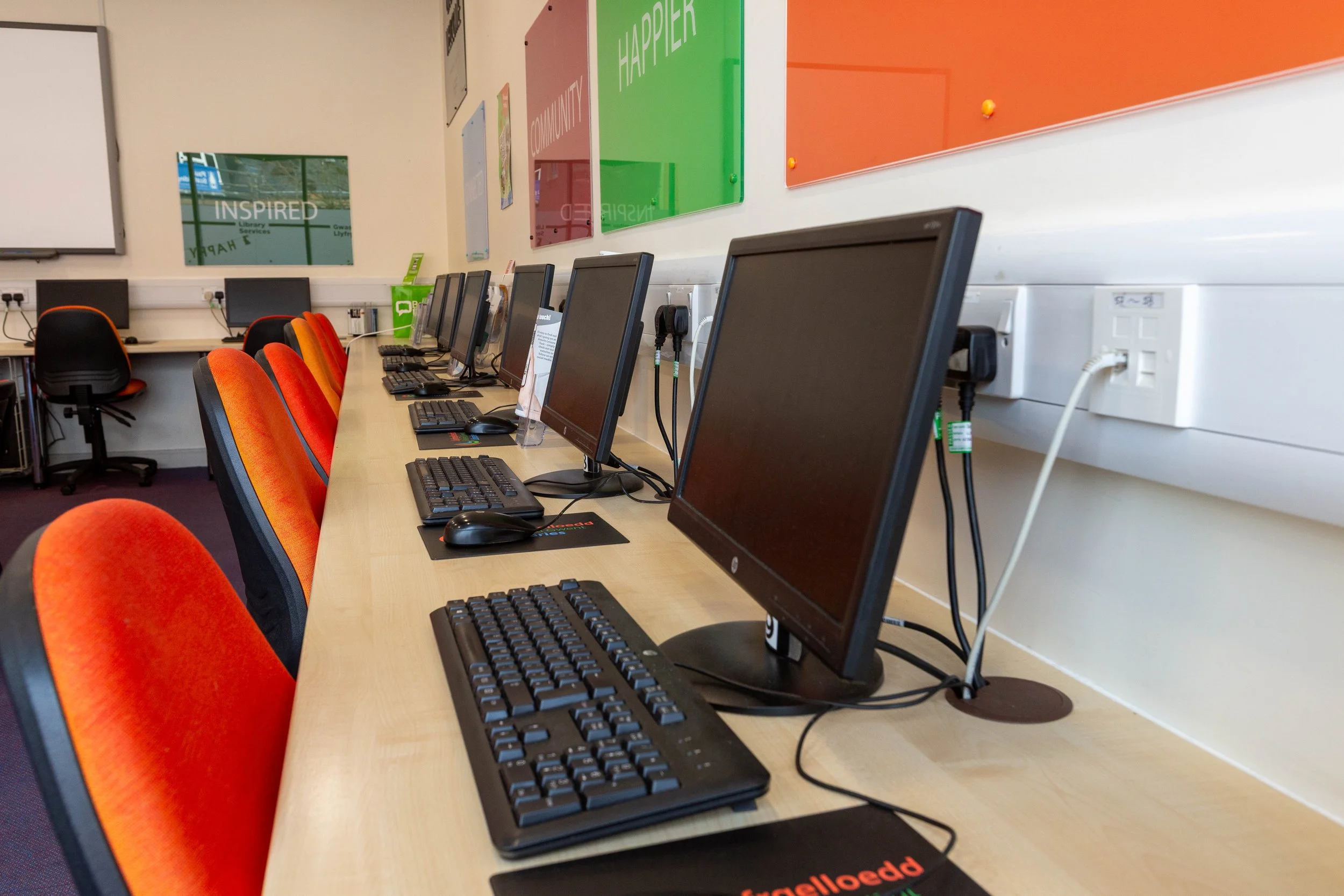 Row of desktop computers with keyboards and mice on a long table in a computer lab, with colorful posters on the wall and chairs with orange and black upholstery.