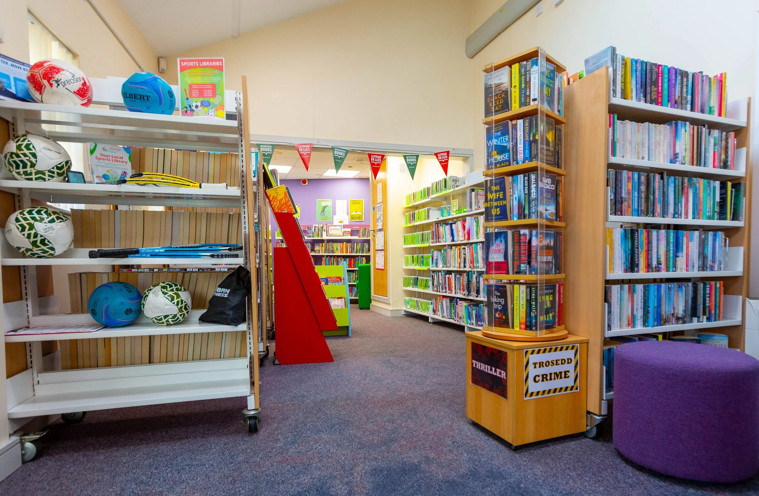 Inside a colorful bookstore with books on shelves, sports equipment on a cart, and traffic light colored bunting banners hanging from the ceiling.