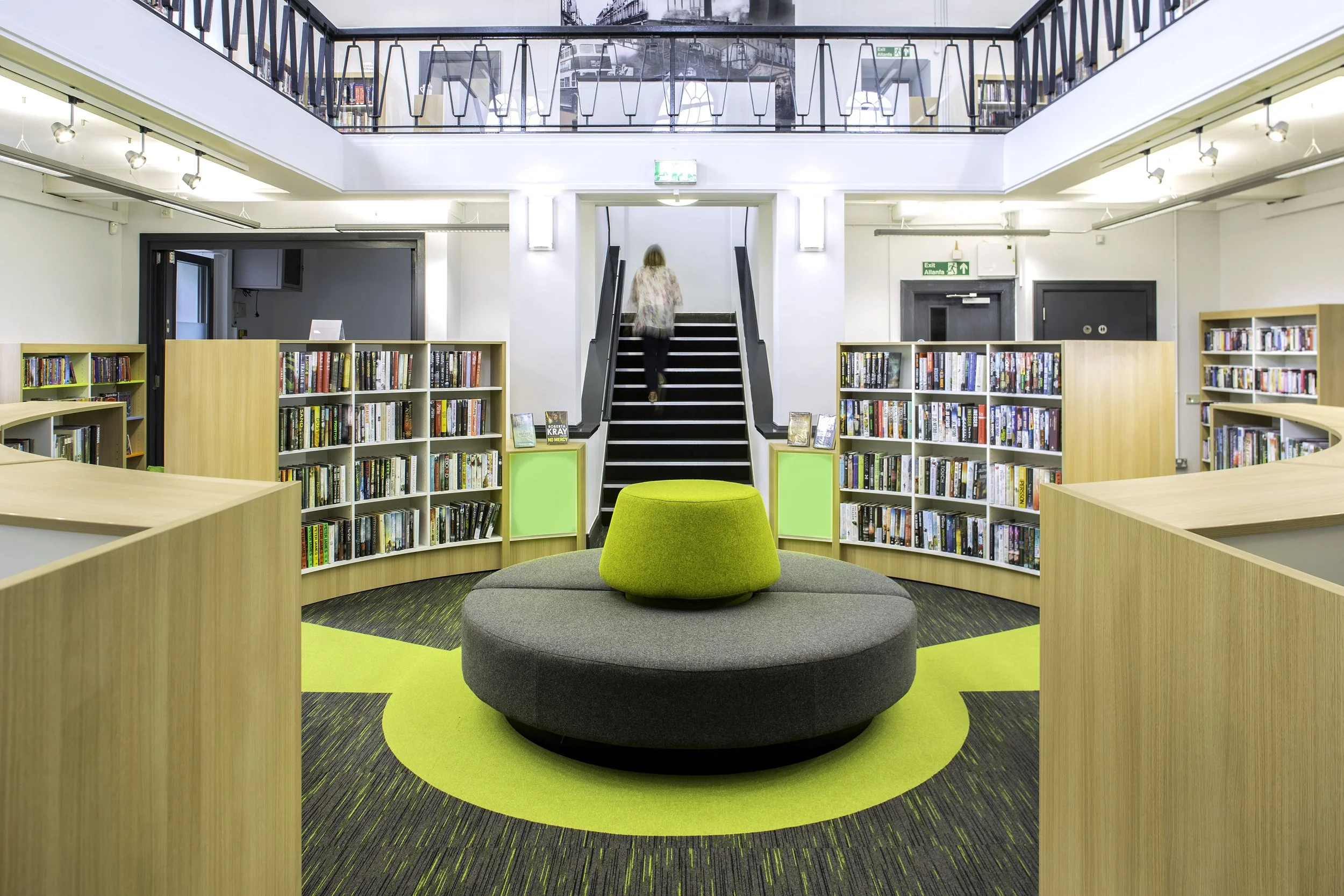 A modern library interior featuring curved bookshelves filled with books, a circular gray and green seating area in the center, and a staircase leading to an upper level with additional bookshelves and seating.
