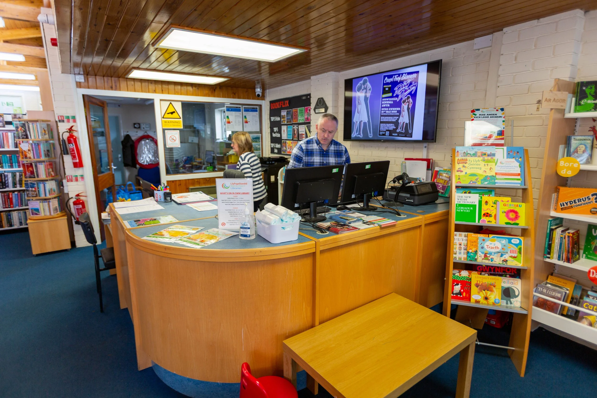 Inside a library with a curved service desk, two staff members working on computers, shelves with children's books, and a large display screen on the wall.
