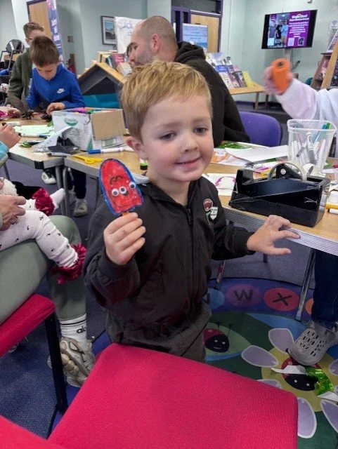 A young boy with red hair and a black jacket sitting at a table in a classroom or library, holding a red craft project with googly eyes. Other children and an adult are visible in the background, along with books and craft supplies on the table.