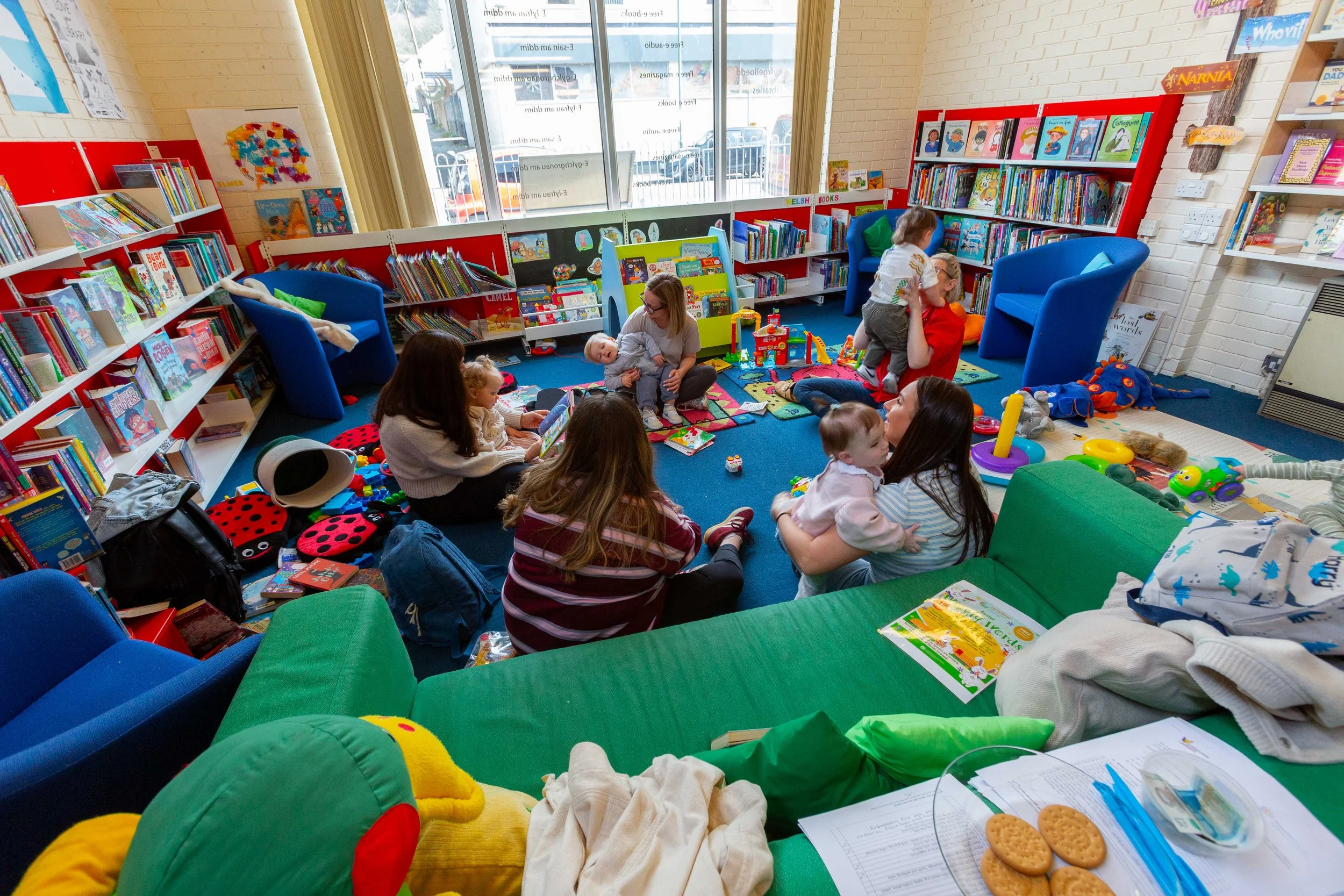 Children and caregivers in a colorful children’s library play and read on the blue carpeted floor, with bookshelves filled with children's books, toys, and colorful furniture surrounding them.