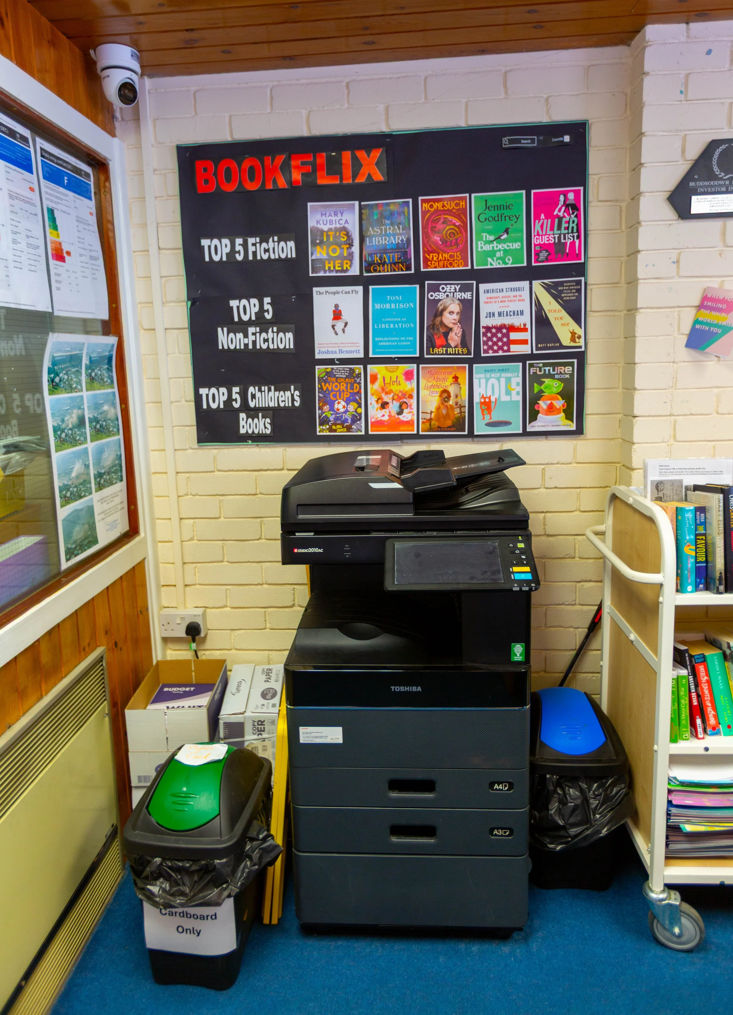 A photo of a library or bookstore corner with a black copier machine, a small bookshelf filled with books, and a wall display listing top 5 fiction, non-fiction, and children's books. There is a recycling bin labeled 'Cardboard Only' and some boxes o