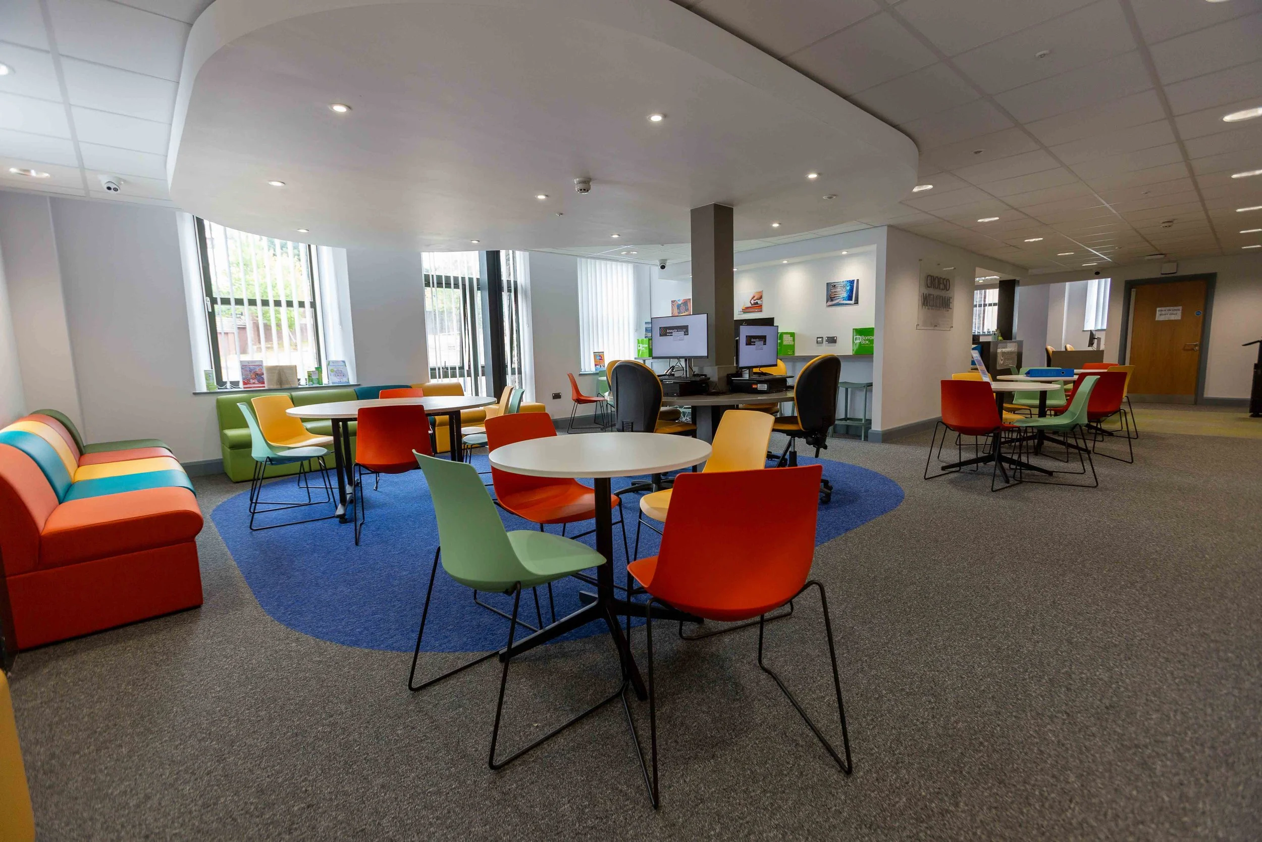 Bright, modern lounge area with colorful chairs around white tables, a blue circular rug, and computer stations in the background with large windows letting in natural light.