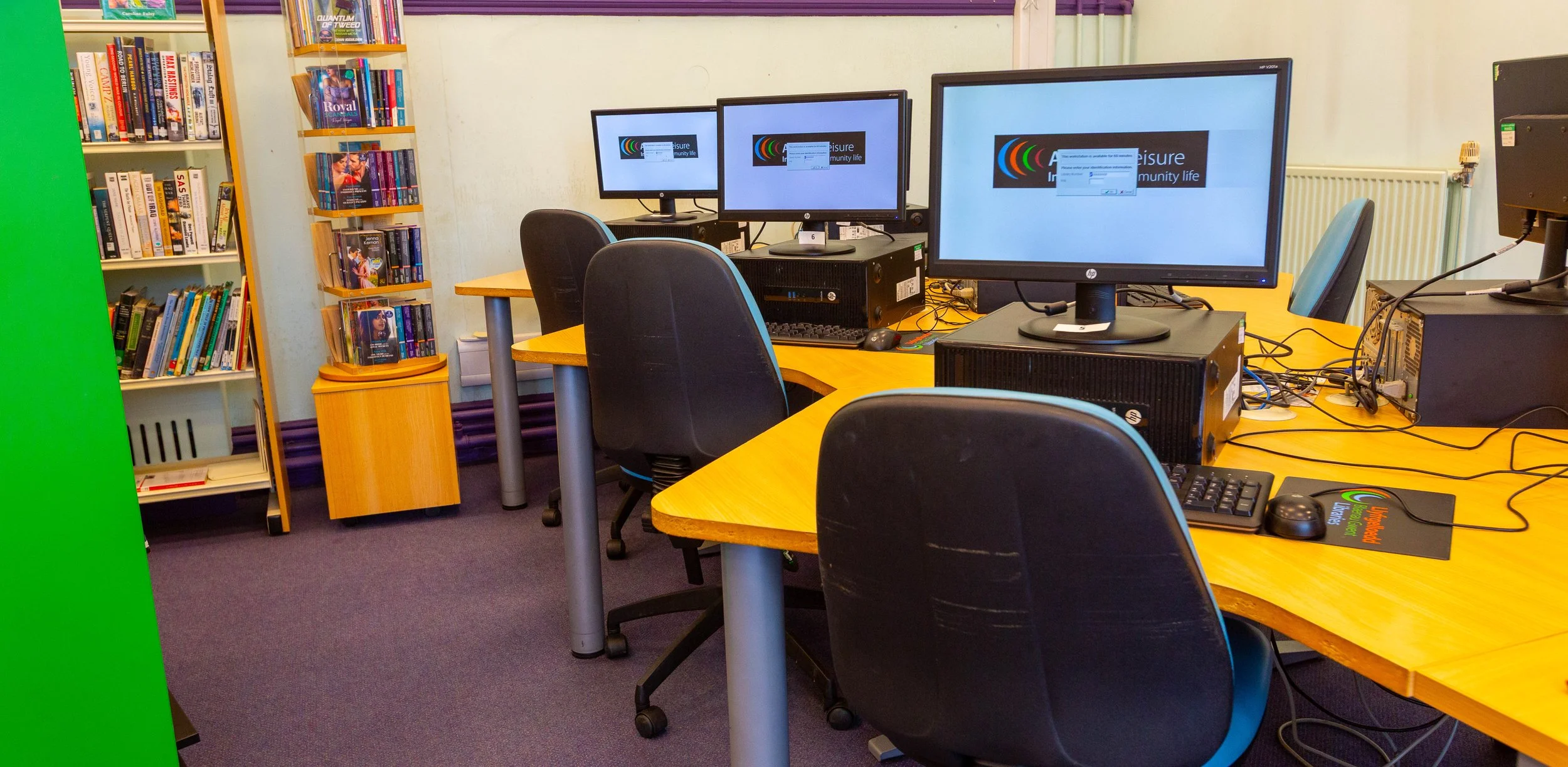 Computer room with three monitors on yellow tables, black chairs, bookshelf with books, and a purple carpet.