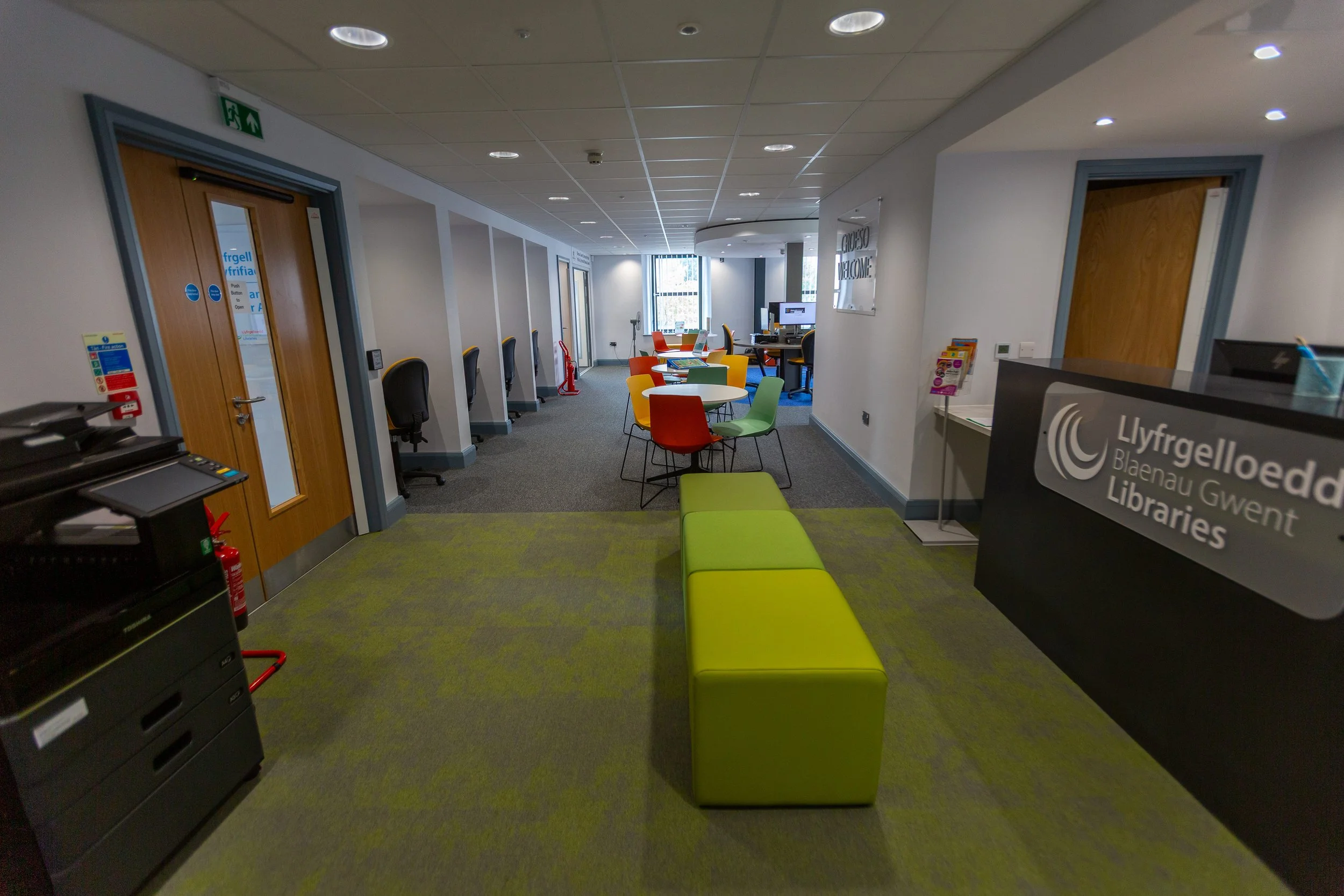 Interior of Blaenau Gwent Libraries with colorful chairs, a green bench, a reception desk, and computer workstations.