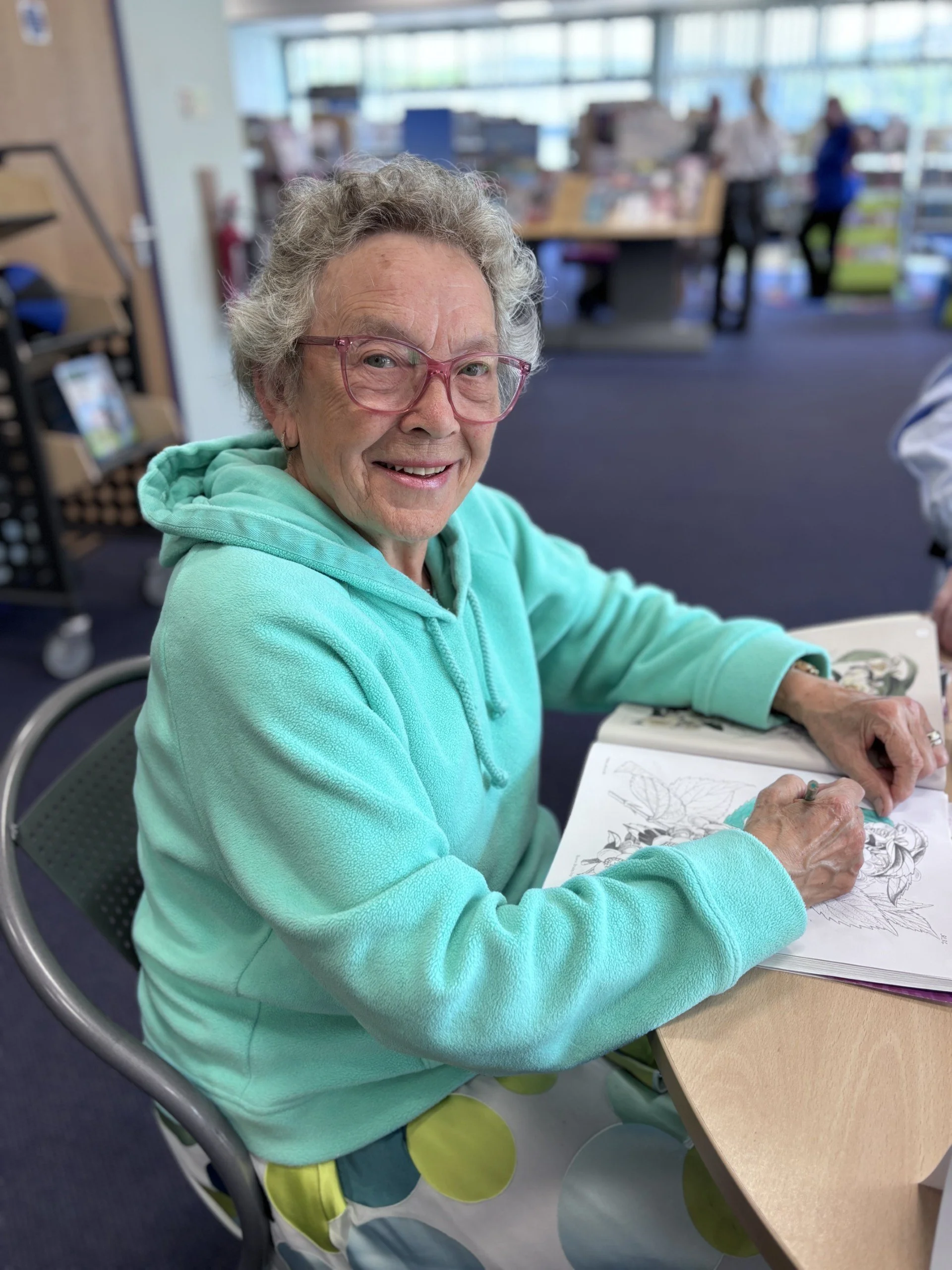 An elderly woman with gray curly hair, glasses, and a bright smile, sitting at a table in a public library or community center, coloring a detailed floral drawing in a coloring book.